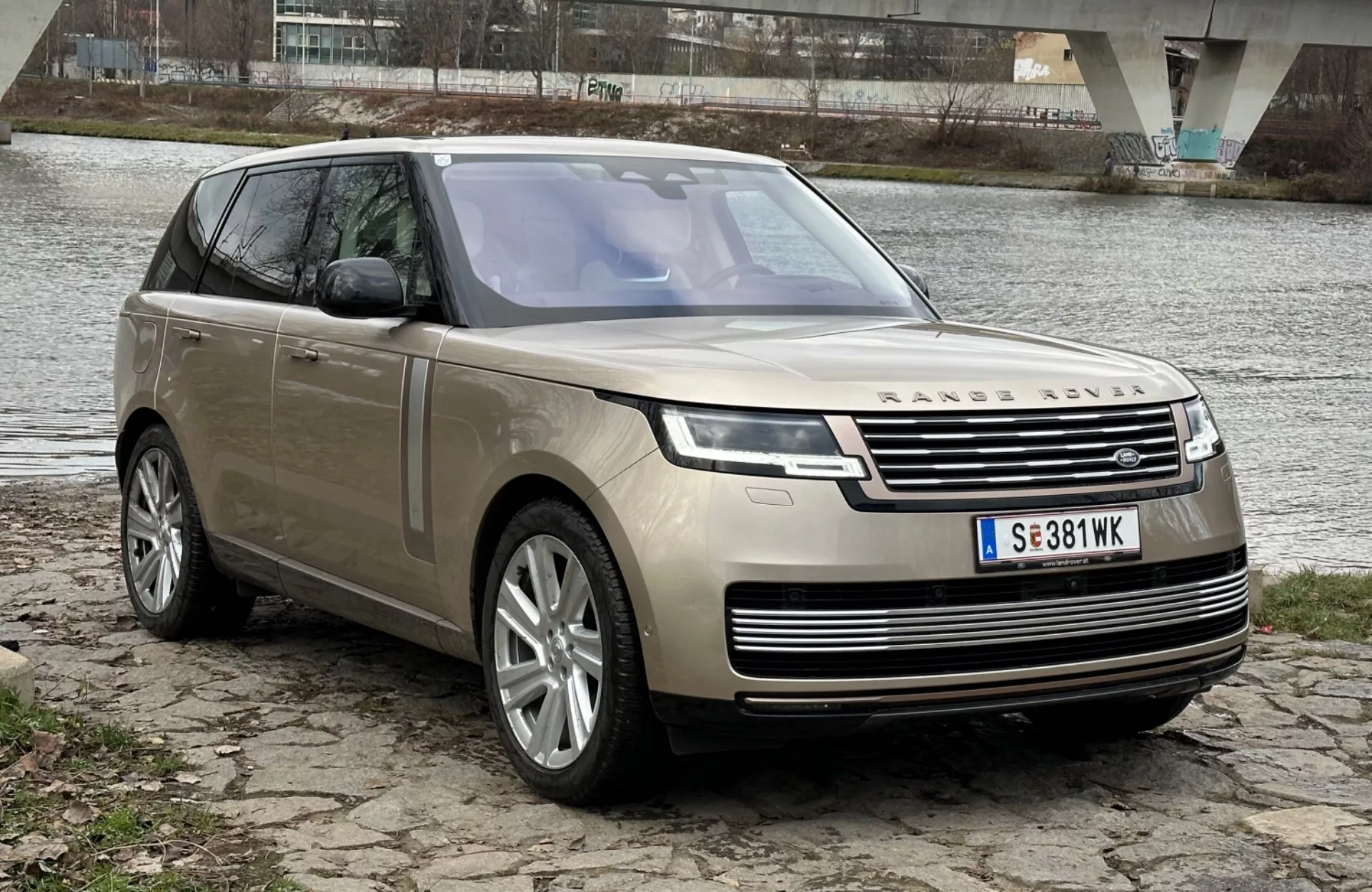Luksusowy SUV Land Rover Range Rover rocznik 2021 zaparkowany na brzegu rzeki z mostem w tle, prezentujący nowoczesny design i cechy technologiczne, takie jak reflektory LED, przedni grill z emblematem, wieloramienne felgi aluminiowe i panoramiczne okno dachowe.