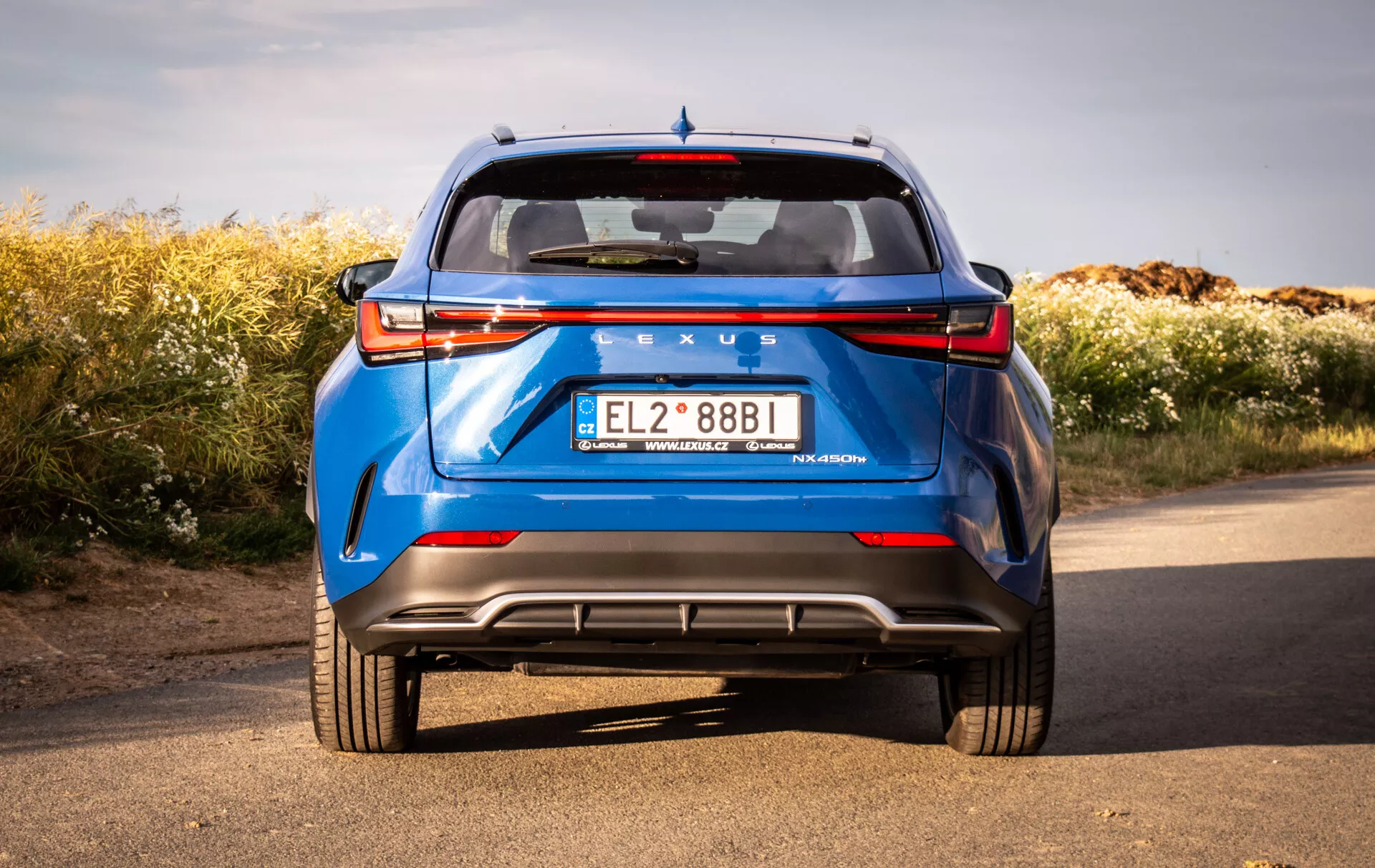 Tylny widok niebieskiego Lexusa NX 2021 z niskiej perspektywy podkreślający solidną budowę i cechy konstrukcyjne, takie jak LEDowe tylne światła, tylna szyba, spojler, wyraźne oznaczenia, tylny zderzak z dyfuzorem, czujniki parkowania, odblaski i podwójny wydech, na tle naturalnym pod bezchmurnym niebem.
