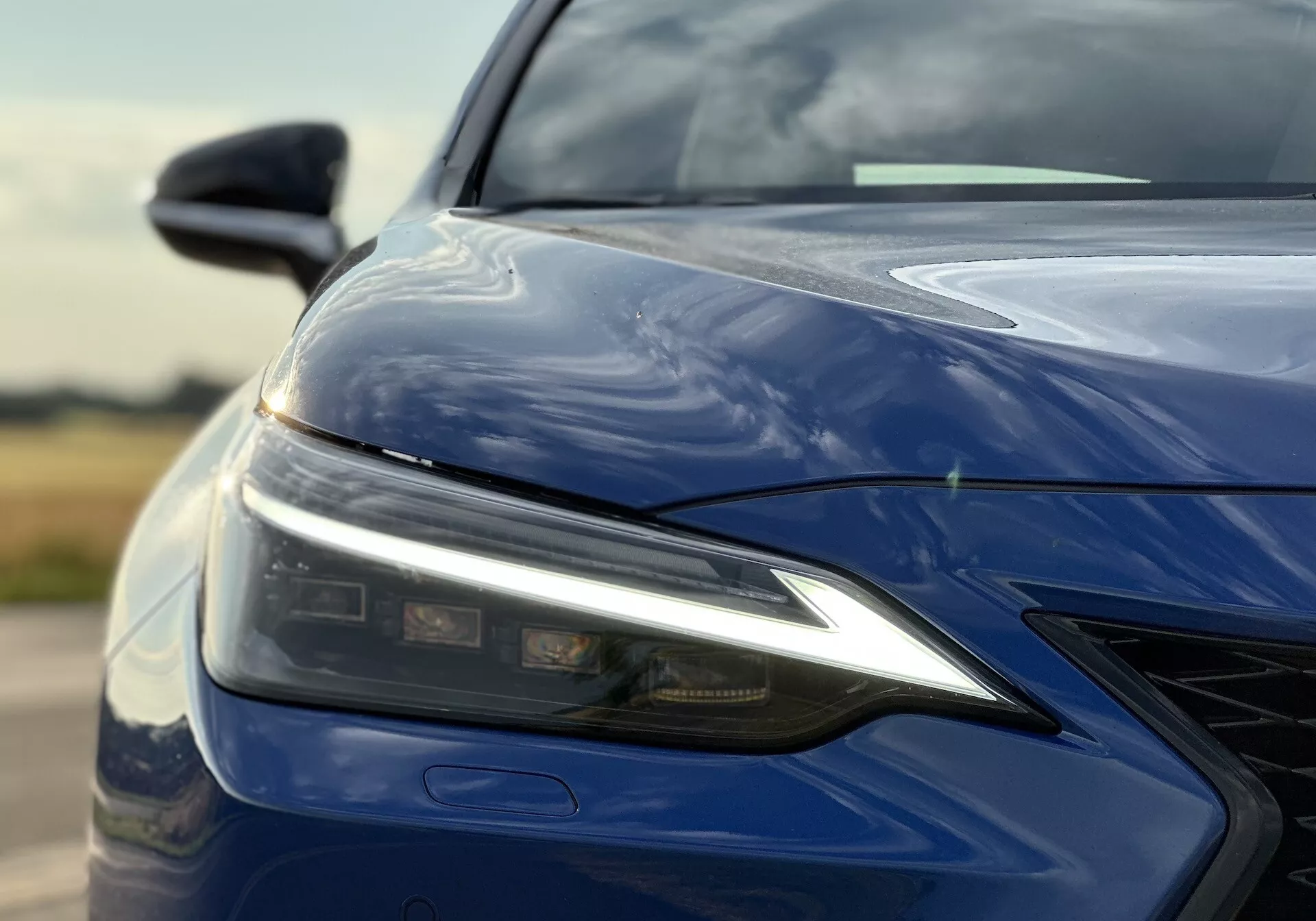 Zbliżenie na lewy przedni kwadrant Lexusa NX 2021 z zespołem oświetlenia LED, rzeźbioną maską, przednim zderzakiem z grillem i błyszczącym, głębokim niebieskim wykończeniem, podkreślając zaawansowaną technologię oświetlenia i aerodynamiczny design.