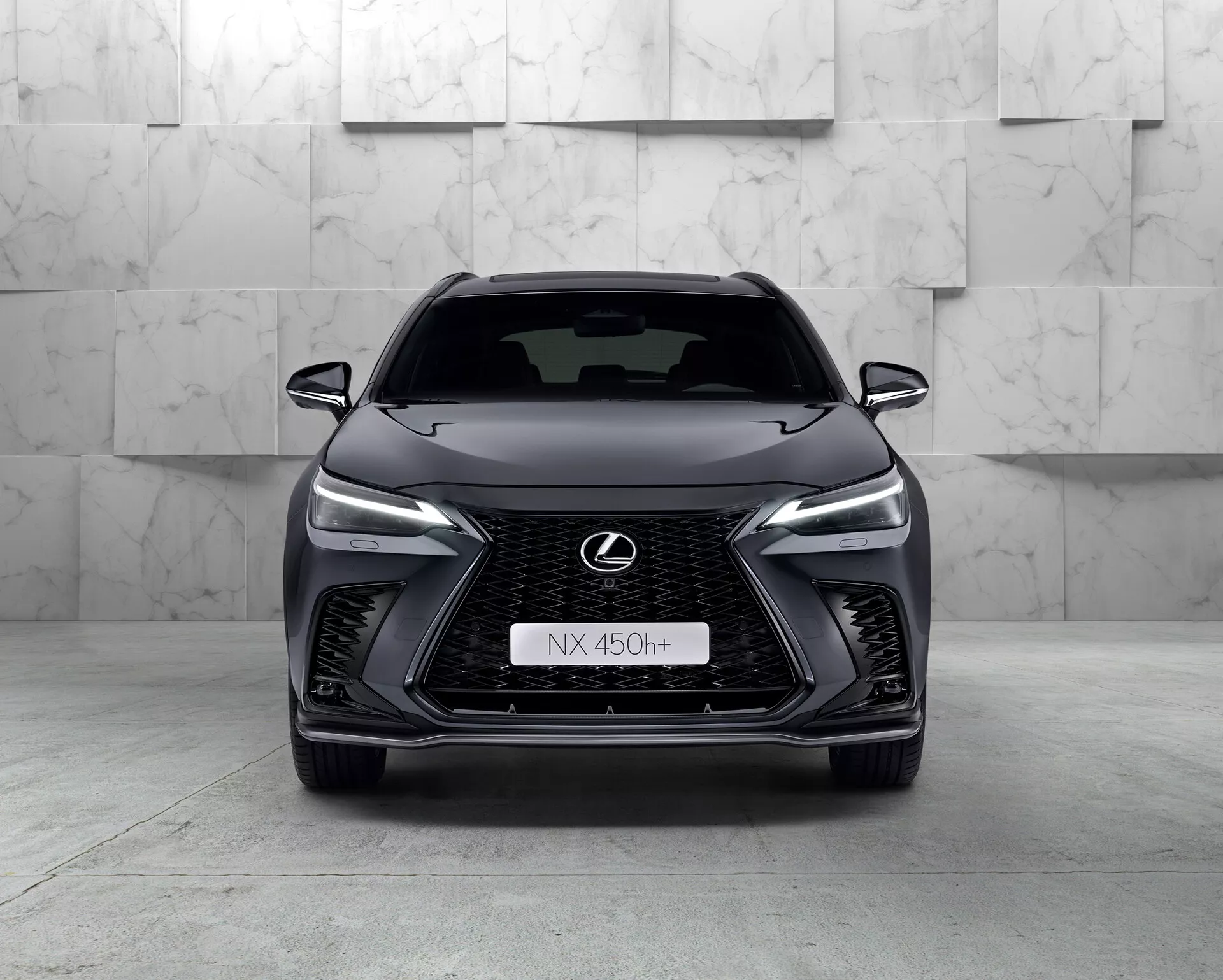 Widok z przodu Lexusa NX 450h+ 2021, eleganckiego nowoczesnego SUV-a prezentującego swoją wyrazistą wrzecionowatą osłonę chłodnicy z siatkowym wzorem, wąskimi reflektorami LED, chromowanym emblematem Lexusa z niebieskim obrysem i agresywnym designem przedniego zderzaka. Hybrydowy charakter samochodu jest oznaczony plakietką 'h+' na grillu. Studio podkreśla krzywizny i cechy pojazdu czystym, kontrolowanym oświetleniem.
