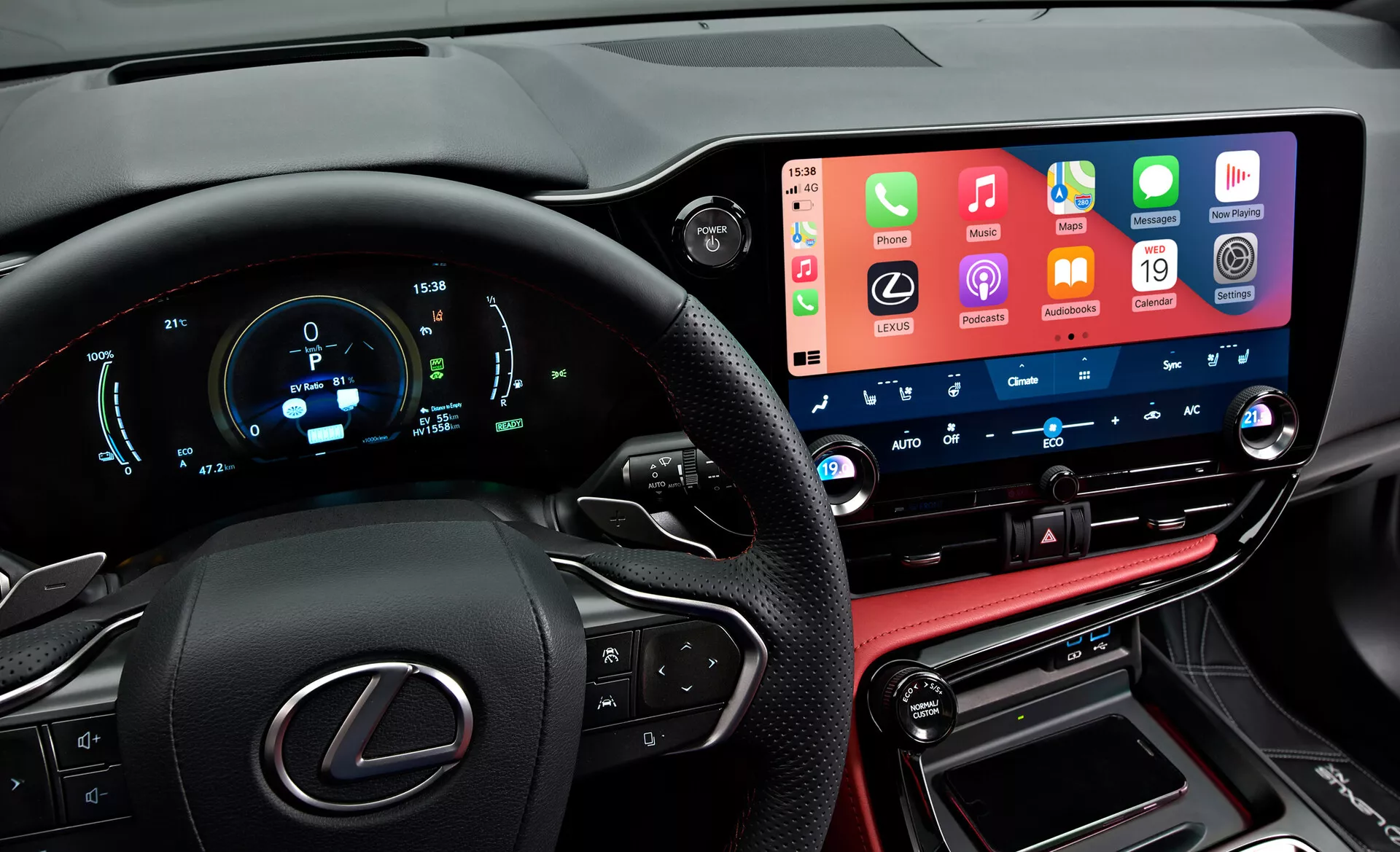 Widok wnętrza Lexus NX 2021, ukazujący nowoczesny, technologicznie zaawansowany kokpit z perspektywy kierowcy. Po lewej widać częściowo logo Lexus na kierownicy z przyciskami multifunkcyjnymi. Cyfrowy zestaw wskaźników wyświetla prędkość, obroty i inne parametry w kolorach niebieskim i białym. Na środku widoczny jest duży ekran dotykowy systemu multimedialnego integrujący funkcje smartfonów. Poniżej znajdują się przyciski i pokrętła do sterowania klimatyzacją z cyfrowymi wyświetlaczami temperatury. Dolna część zawiera konsolę szytą skórą z dźwignią zmiany biegów oraz portami komunikacyjnymi, odzwierciedlając luksusowe wykończenia pojazdu.