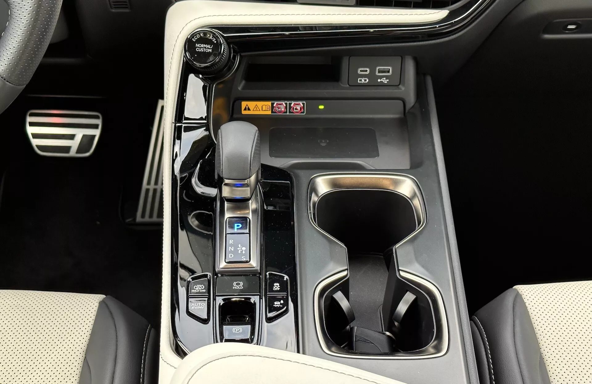 Widok konsoli środkowej Lexusa NX 2021 pokazujący automatyczną dźwignię zmiany biegów z kożuchem, selektor trybu jazdy, uchwyty na kubki, przyciski sterowania pojazdem, potencjalne skróty do systemu informacyjno-rozrywkowego i elementy luksusowego designu wnętrza