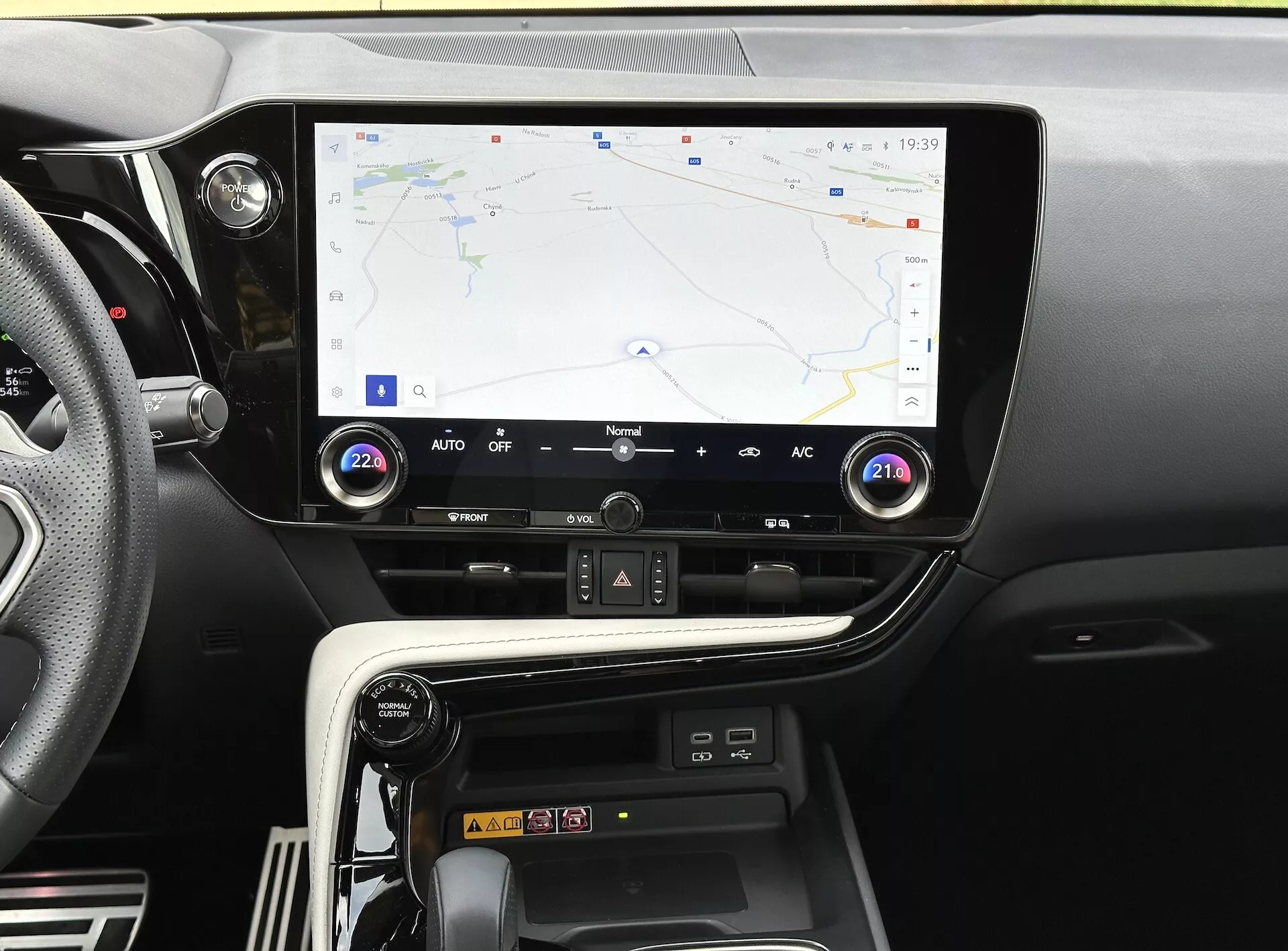 Konsola środkowa Lexusa NX 2021 z ekranem dotykowym wyświetlającym nawigację GPS, przyciski i pokrętła kontroli klimatyzacji oraz przegródkę na ładowanie smartfona