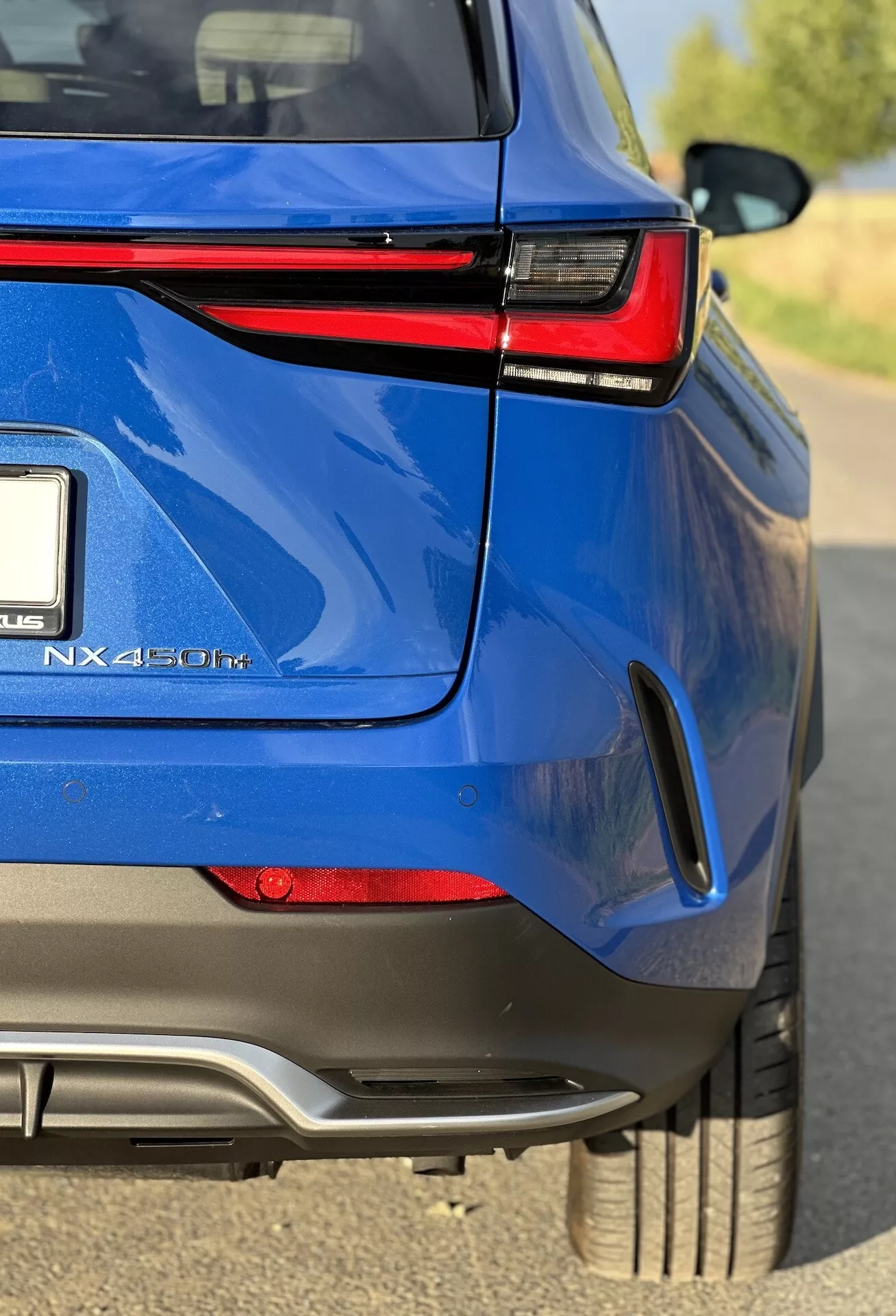 Zbliżony ujęcie pod kątem tylnej części niebieskiego hybrydowego SUV-a Lexus NX 450h+, podkreślające połyskliwy wykończenie, LED-owe tylne światła, emblemat hybrydowy i sportowe cechy stylistyczne