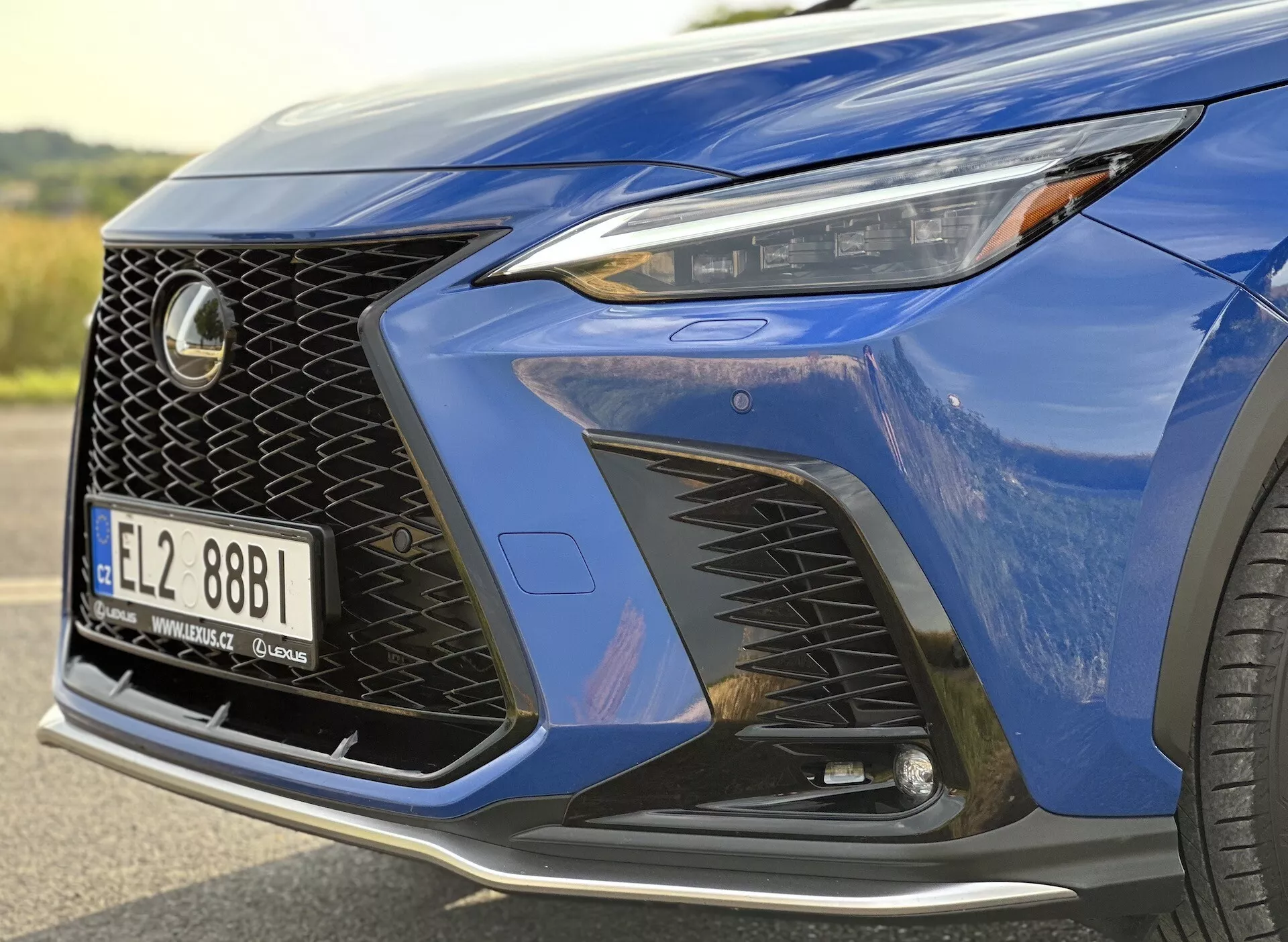 Zbliżenie na przedni róg Lexusa NX 2021 z reflektorem LED, charakterystycznym wzorem atrapy chłodnicy oraz aerodynamicznym przednim zderzakiem z dużymi wlotami powietrza, podkreślone metalicznym, głębokim niebieskim lakierem i widokiem na luksusowe koło ze stopu.