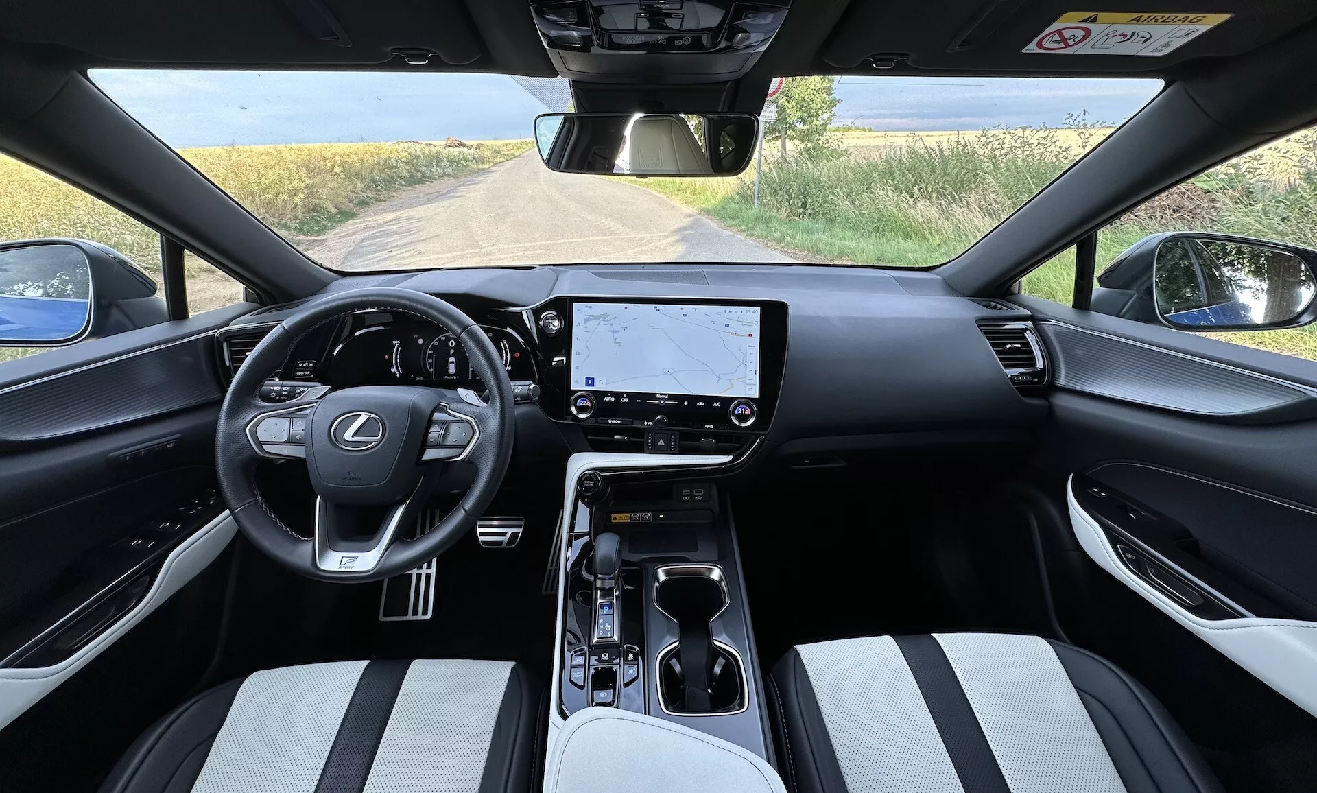 Widok wnętrza Lexusa NX 2021 z perspektywy kierowcy, przedstawiający centralnie umieszczoną kierownicę z logo Lexusa i przyciskami do sterowania, duży ekran dotykowy systemu infotainment na desce rozdzielczej, minimalistyczne kontrolki klimatyzacji, selektor biegów automatycznej skrzyni biegów na konsoli środkowej z uchwytami na napoje i schowkiem, tapicerowane dwukolorowym materiałem siedzenia, oraz częściowy widok na wiejski krajobraz przez przednią szybę.