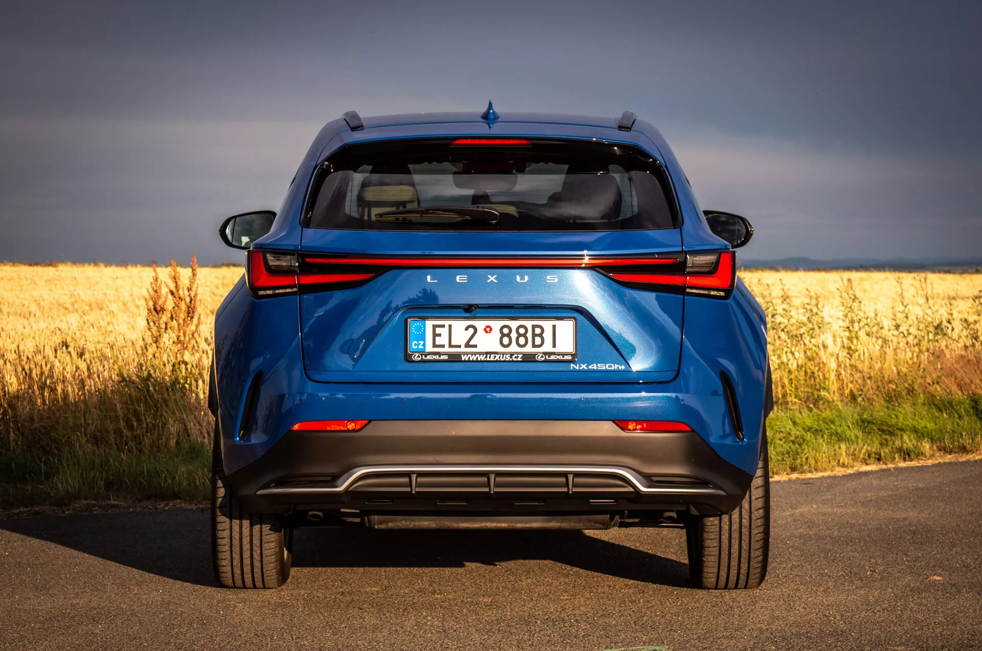 Tylny widok na niebieskie SUV Lexus NX 2021 na asfaltowej drodze z złotym polem w tle, podkreślający technologię hybrydową i cechy projektowe