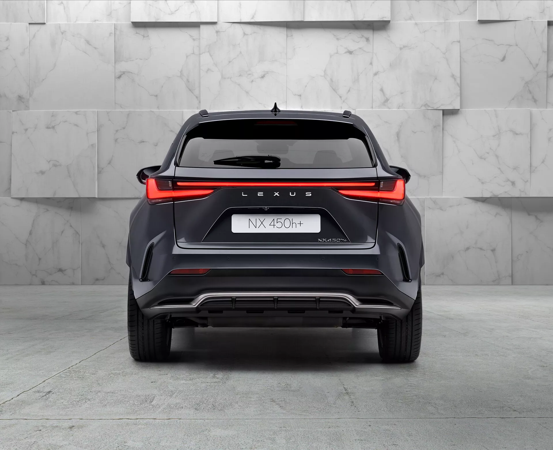 Tylny widok na hybrydowego SUV-a Lexus NX 450h, uchwycony z niskiego kąta lekko przekrzywionego w lewo, ukazujący pełnowymiarowy wzór tylnego światła, markę 'LEXUS', oznaczenie modelu 'NX 450h', nazwę 'NEXUS' i symbol hybrydy 'h'. Zderzak o kanciastych elementach stylizacji, z cechami wentylacyjnymi i srebrną osłoną, widocznym podwójnym wylotem wydechu po lewej stronie, wszystko w środowisku showroomu z wypolerowaną podłogą i neutralnym tłem.