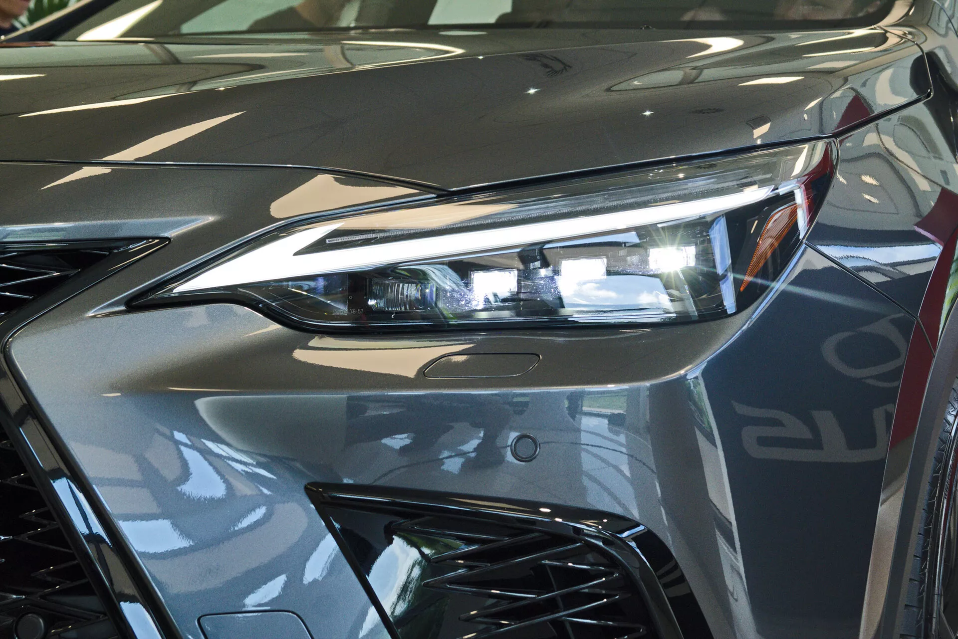 Zbliżenie przedniej części Lexusa NX 2021, skupiające się na zespole reflektorów po stronie kierowcy z zaawansowaną technologią oświetlenia LED i części agresywnie stylizowanej kratki wlotu powietrza. Obraz uchwyca odbijający blask metalicznego srebrnego lakieru i dynamiczne linie przyczyniające się do aerodynamicznej estetyki pojazdu. Fotografowane w pomieszczeniu, prawdopodobnie w salonie wystawowym, podkreślając luksusowy wygląd i wysoką jakość wykończenia pojazdu.