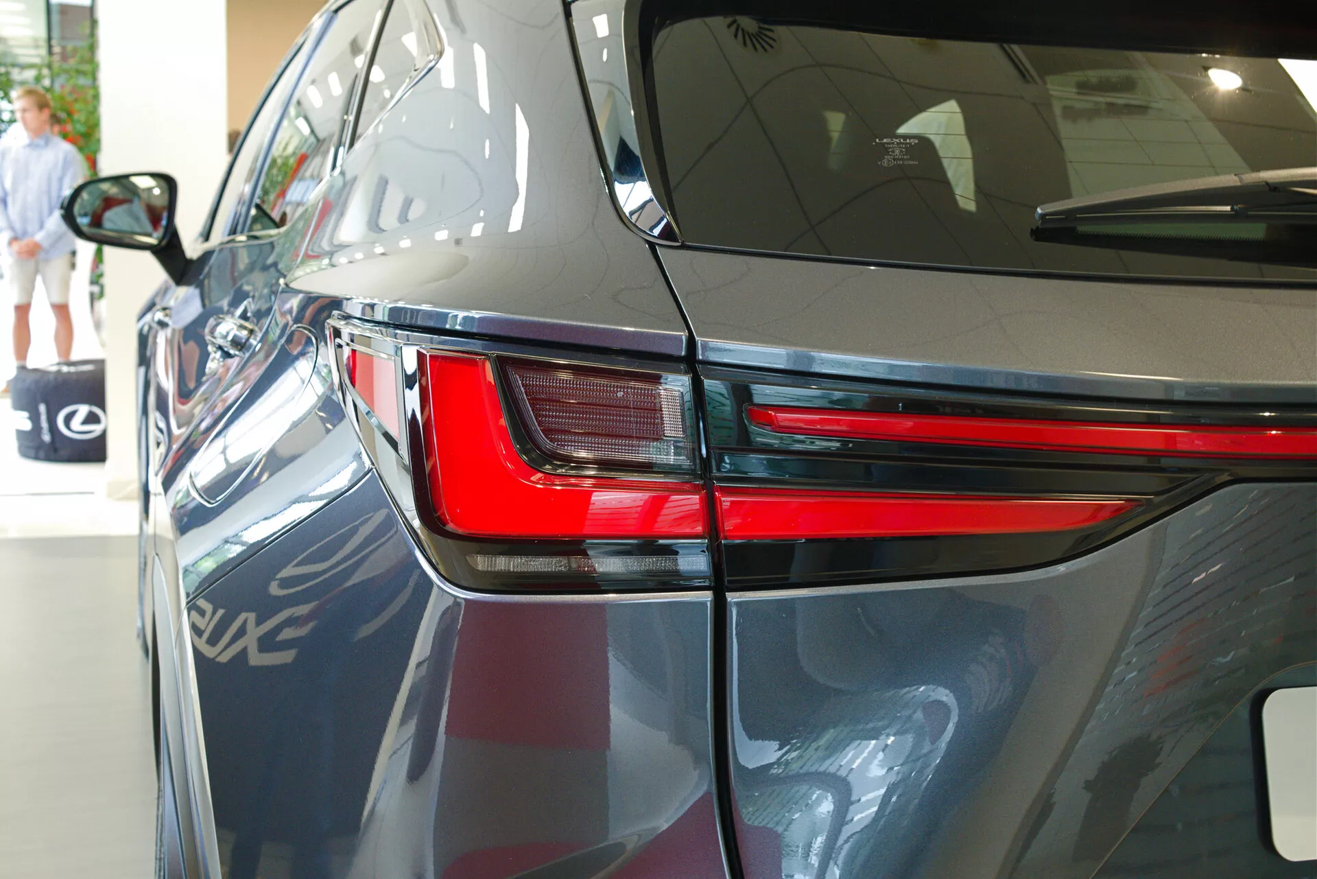 Zbliżenie Lexusa NX 2021 z tyłu w trzech czwartych pod kątem, podkreślające elegancki design tylnego światła LED, część tylnego panelu i fragment pokrywy bagażnika. Obraz pokazuje ciemny, błyszczący lakier samochodu, aerodynamiczne linie nadwozia i sugeruje wewnętrzne środowisko wystawowe z odbiciami innego pojazdu, elementów architektonicznych i osoby idącej w tle.