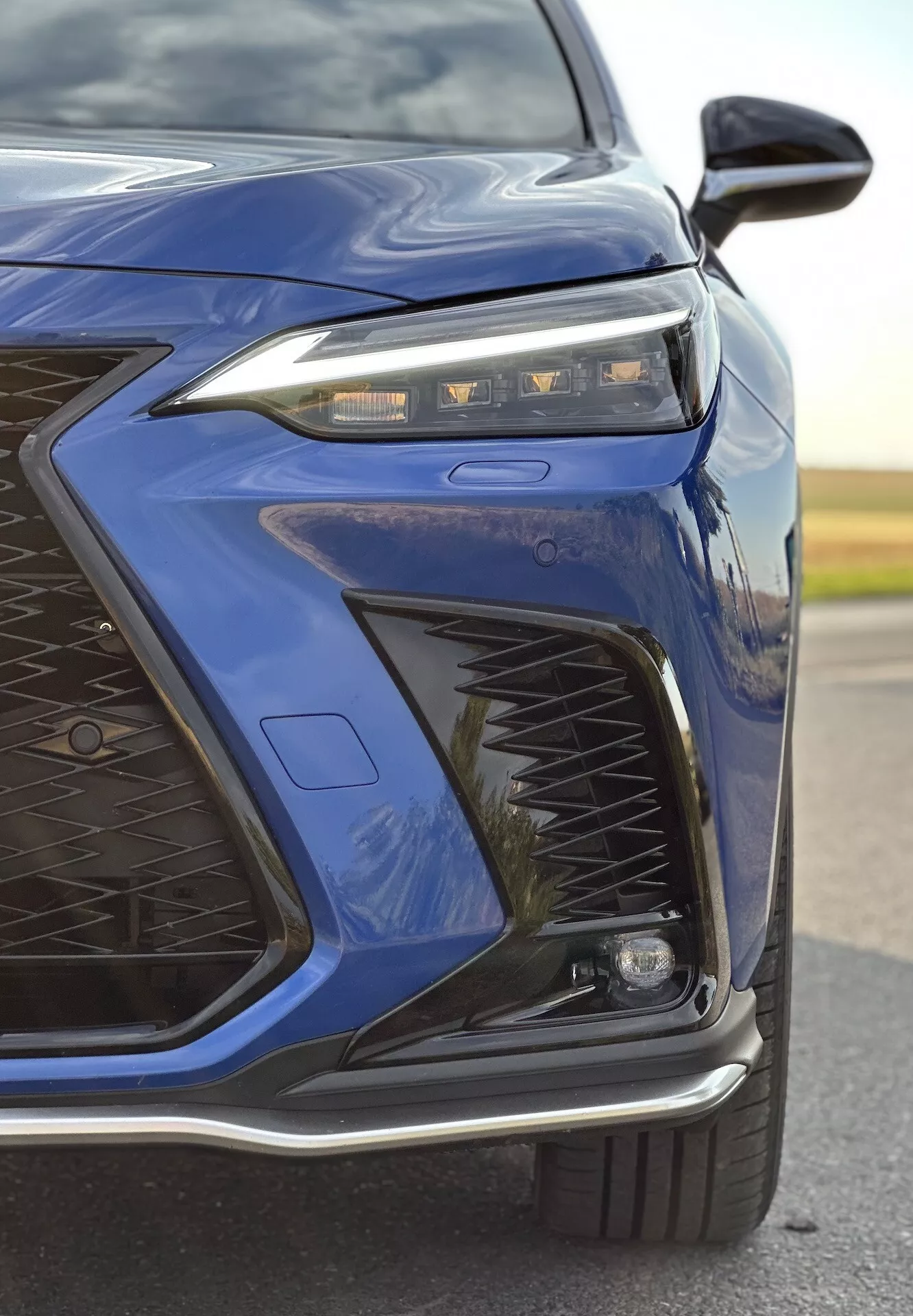 Zbliżenie na przedni lewy róg Lexusa NX z 2021 roku w ciemnoniebieskim kolorze, podkreślające zaawansowaną konstrukcję reflektorów LED, skomplikowany wzór grilla, sportowy przedni zderzak oraz potencjalne funkcje adaptacyjnego oświetlenia i aerodynamiczne z połyskującym metalicznym wykończeniem