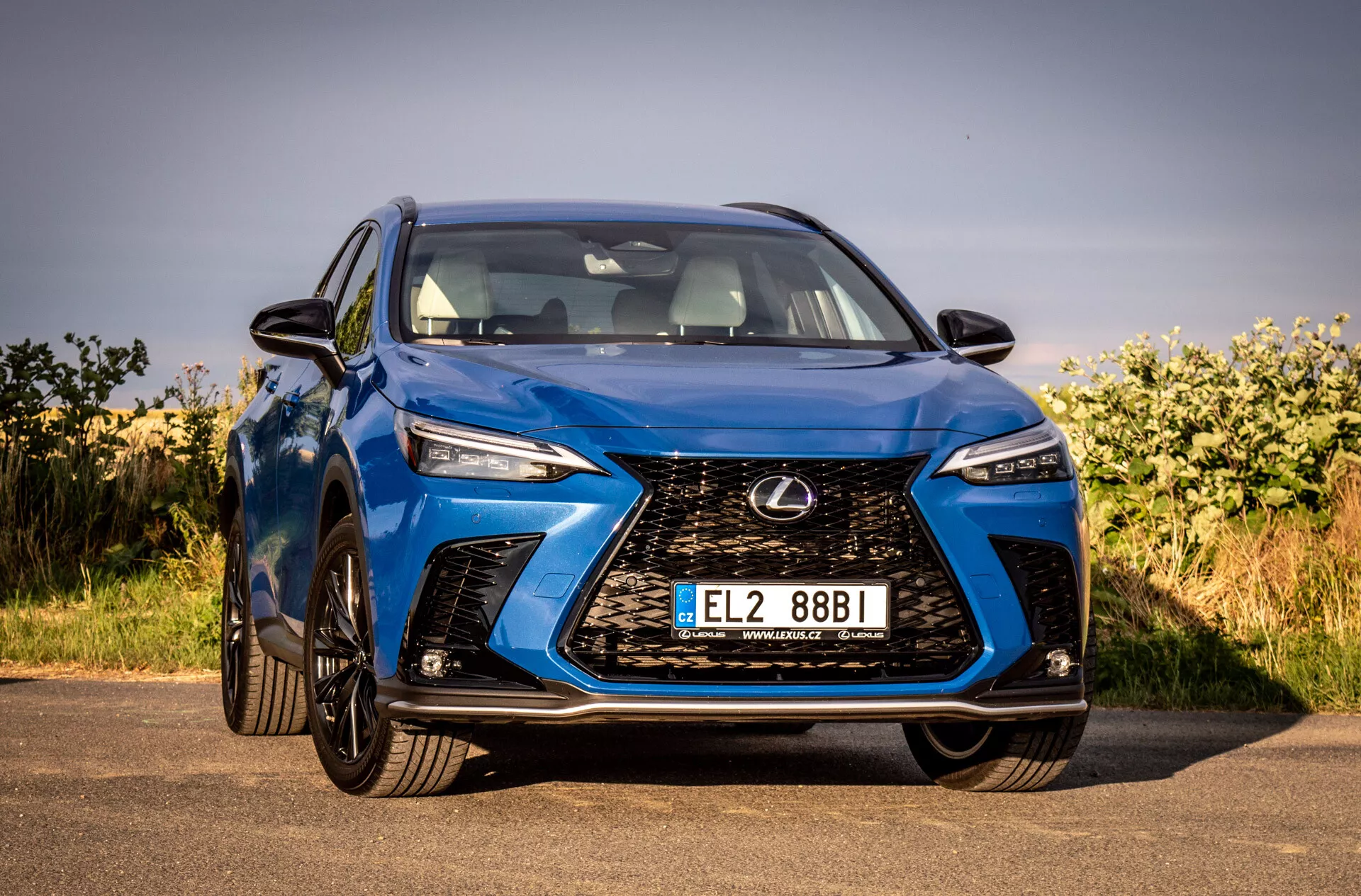 Widok z przodu na niebieskie SUV Lexus NX 2021 podkreślający jego skomplikowaną kratkę chłodnicy typu spindle, kanciaste reflektory LED, błyszczące wykończenie i aerodynamiczny design, zaparkowany na zewnątrz pod bezchmurnym niebem