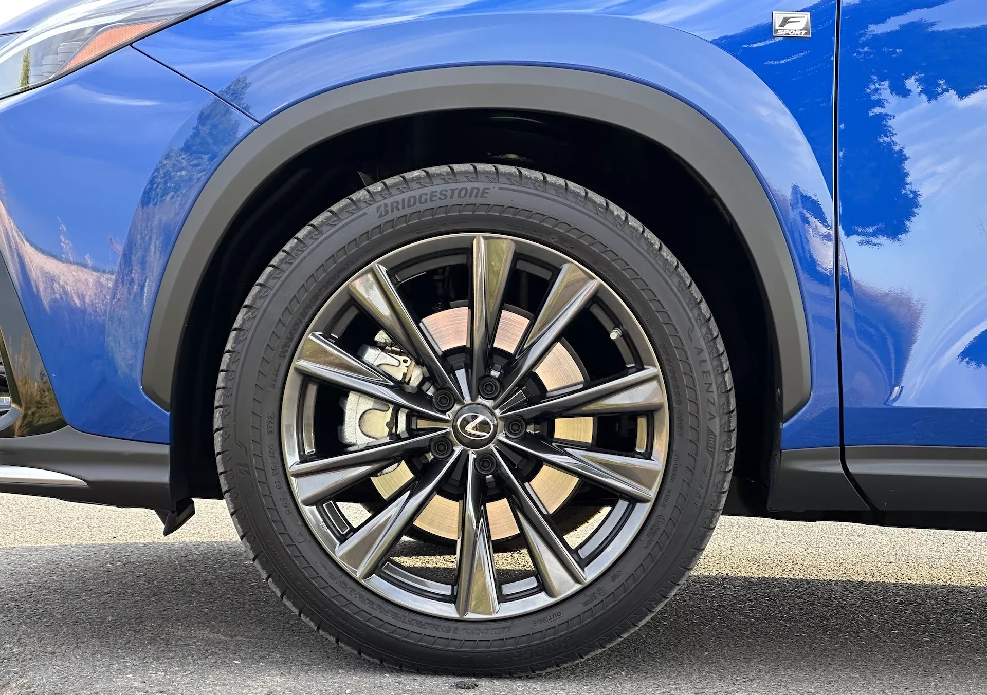 Szczegółowy widok przedniego koła po stronie pasażera Lexusa NX 2021 z wieloramieniowym wzorem o dwutonowej wykończeniu i oponą Bridgestone, na tle żywego niebieskiego lakieru