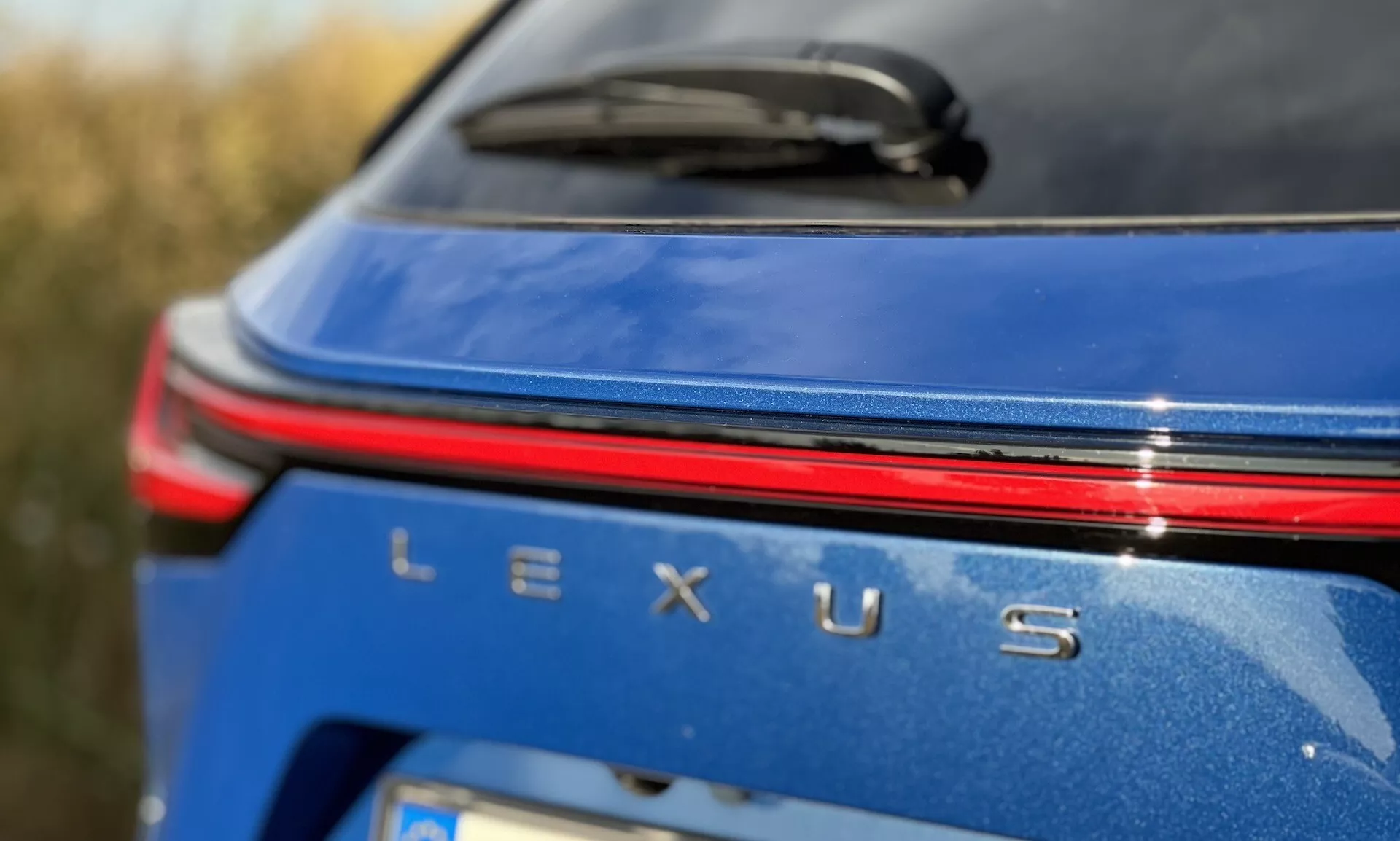 Zbliżenie na tylny obszar brandingu Lexusa NX 2021 z srebrną odznaką Lexusa, czerwonym paskiem świetlnym nad nazwą, na metalicznie niebieskim nadwoziu w warunkach dziennego światła