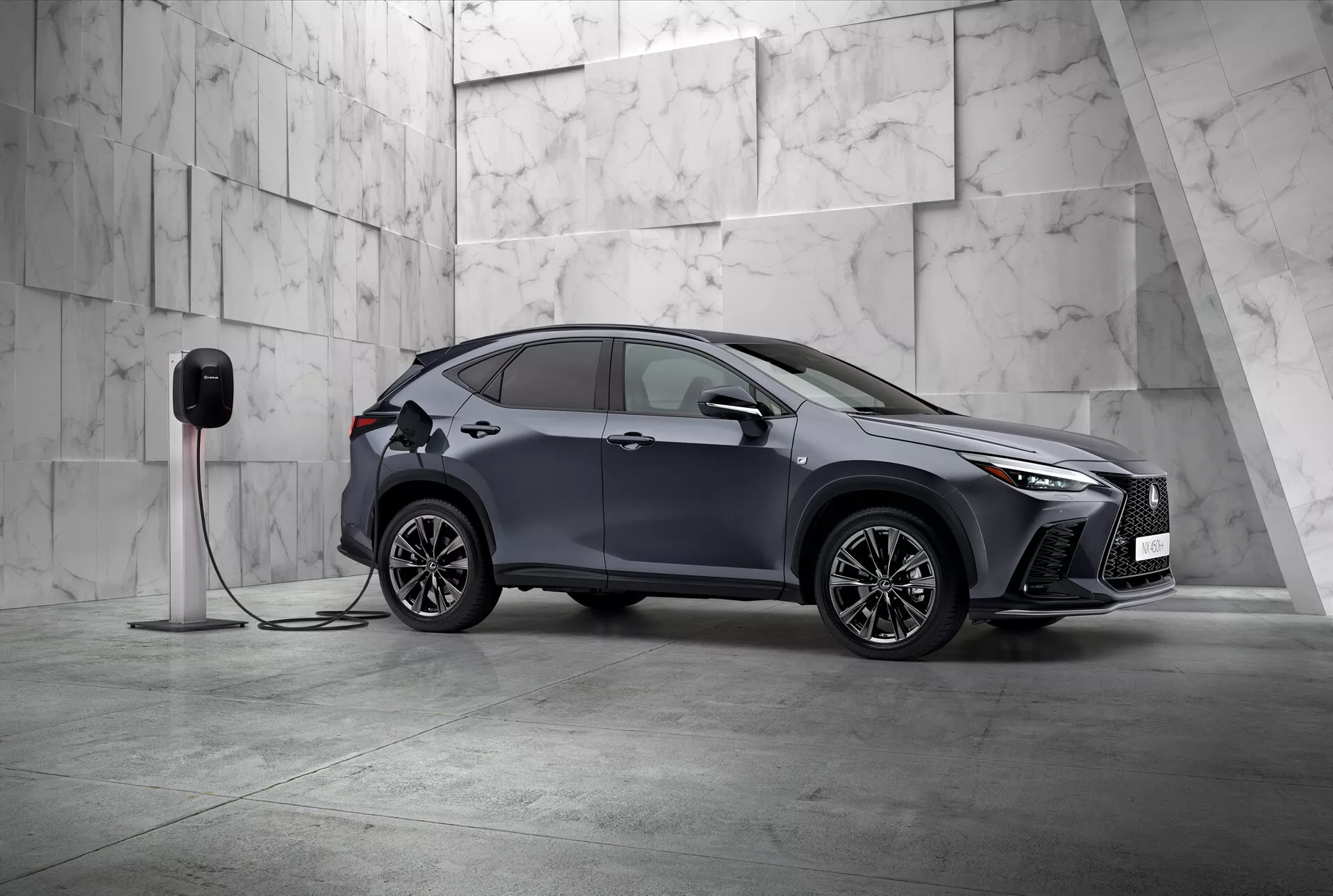 Obraz przedstawia 2021 Lexus NX, nowoczesny kompaktowy SUV o eleganckim i sportowym designie, sfotografowany z perspektywy trzech czwartych z przodu. Widoczne są lewa strona pojazdu i częściowo jego przód, ustawione ukośnie w minimalistycznym otoczeniu ozdobionym dużymi jasnymi marmurowymi płytkami, sugerującymi atmosferę premium. Lexus NX posiada charakterystyczne kanciaste reflektory LED i wyraźny przedni grill, podkreślając swoją wyrafinowaną naturę. Widoczne są aerodynamiczne lusterka boczne, skomplikowane felgi ze stopów wieloramiennych oraz wpuszczane klamki drzwi, które podkreślają sportowy i zaawansowany design. Ważnym detalem jest podłączenie auta do stacji ładowania elektrycznego, co wskazuje, że jest to pojazd elektryczny lub hybryda plug-in. Przedni błotnik obejmuje port ładowania, wskazując na elektryczne możliwości. Ten statyczny obraz skupia się na przedstawieniu designu i funkcji technologicznych pojazdu, w spokojnej, zrównoważonej kompozycji z dużą ilością negatywnego przestrzeni, przykładnie ekskluzywność i innowacje w designie motoryzacyjnym.