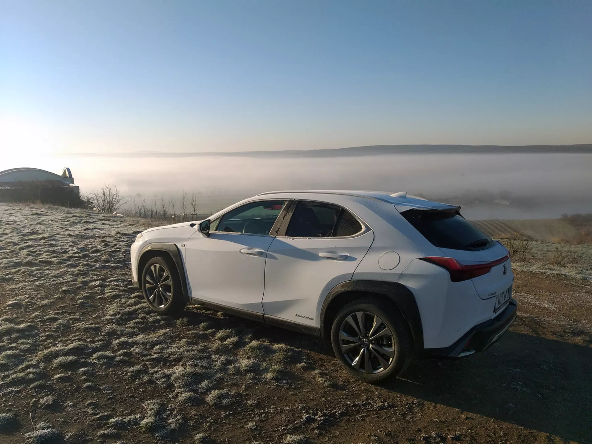 Crossover SUV Lexus UX 2019 zaparkowany na trawie pokrytej szronem z tłem mglistej krajobrazu, przedstawiający profil pojazdu z tylnego trzykwaterowego kąta podkreślającego boczne linie charakterystyczne, łuki kół, zwężający się dach i felgi aluminiowe.