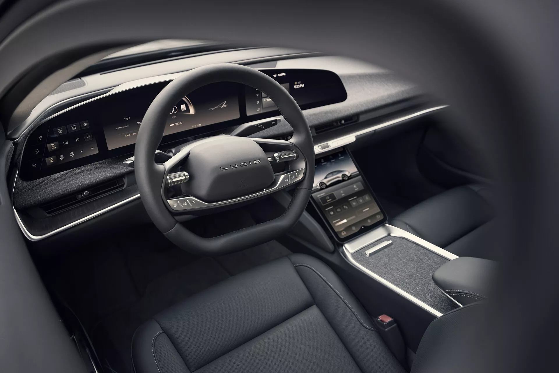 Zbliżenie na kokpit kierowcy Lucid Air 2022 z perspektywy tylnego siedzenia, ukazujące nowoczesną wielofunkcyjną kierownicę z przyciskami sterowania, szeroki cyfrowy wyświetlacz deski rozdzielczej, pionowo zorientowany centralny ekran dotykowy z otworami wentylacyjnymi, elegancką konsolę środkową z minimalną ilością fizycznych przycisków i luksusowe wnętrze z ciemną kolorystyką zawierającą materiały wysokiej jakości.