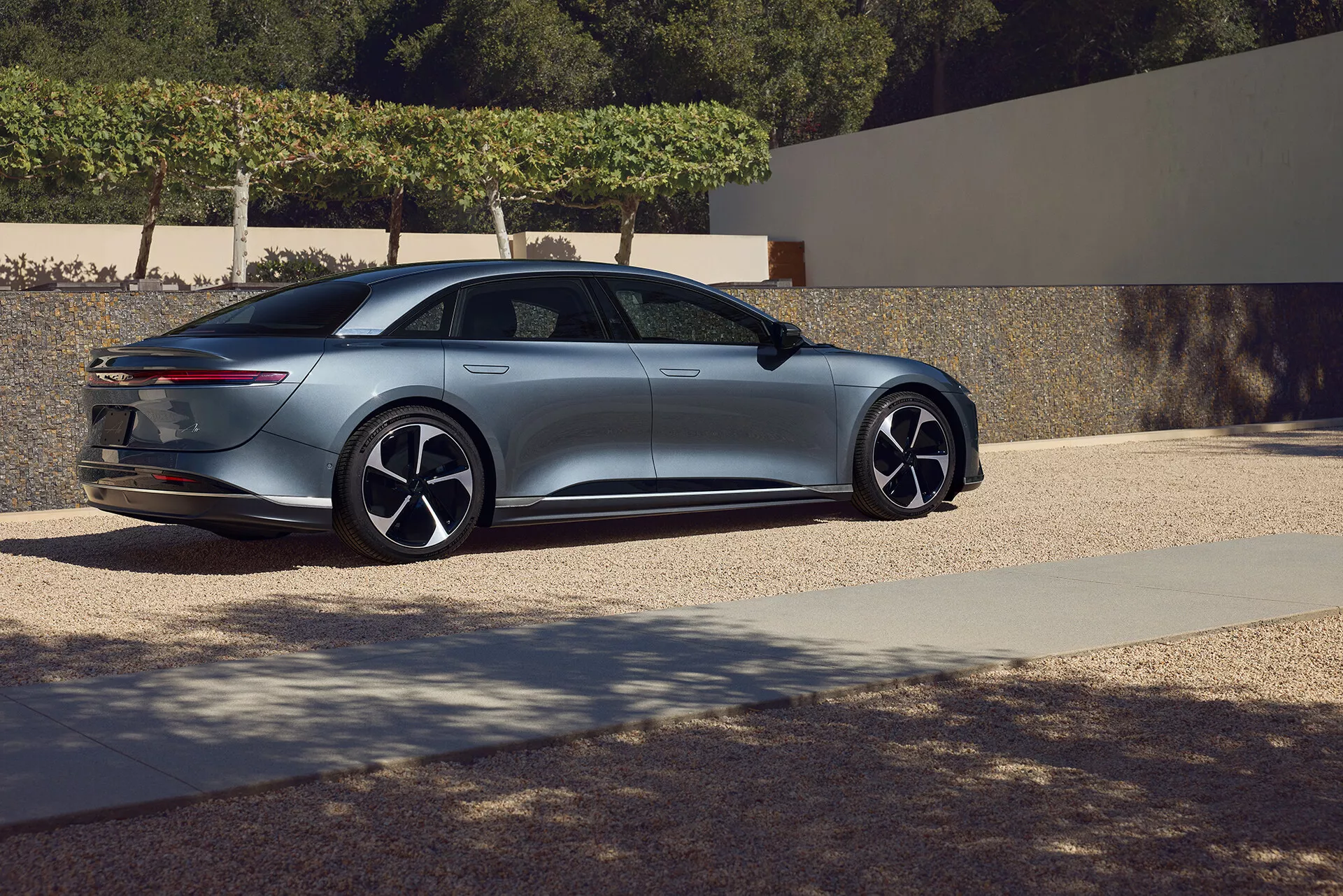 Trzy-czwartowy widok boczny od tyłu sedana Lucid Air 2022 pierwszej generacji. Ujęcie pod niskim kątem podkreśla elegancki i aerodynamiczny design pojazdu, z płynną linią dachu przechodzącą w bagażnik w stylu fastback z wąskimi, poziomo zorientowanymi światłami tylnymi. Profil boczny pokazuje duże, wieloszprychowe felgi i wpuszczane klamki drzwi. Smukłe lusterka boczne i opadający przód sugerują zaawansowane funkcje technologiczne, wzbogacając luksusowy i nowoczesny wygląd samochodu. Sceneria wydaje się być rezydencjalnym obszarem, co podkreśla estetykę premium.