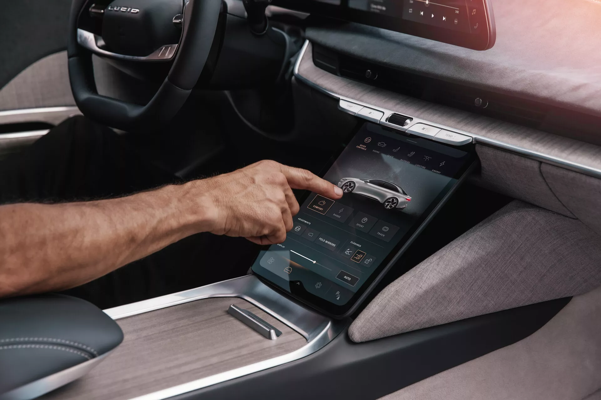Szczegółowy widok konsoli środkowej w Lucid Air 2022 pierwszej generacji pod kątem ukośnym, podkreślający pionowo zorientowany ekran dotykowy, z którym współpracuje prawa ręka osoby, ilustrując zaawansowany system informacyjno-rozrywkowy z różnymi funkcjonalnymi kontrolkami i kombinacją tekstylnych i metalowych paneli, sugerujący nowoczesny, ekskluzywny wystrój wnętrza skoncentrowany na zaawansowanej technologii.