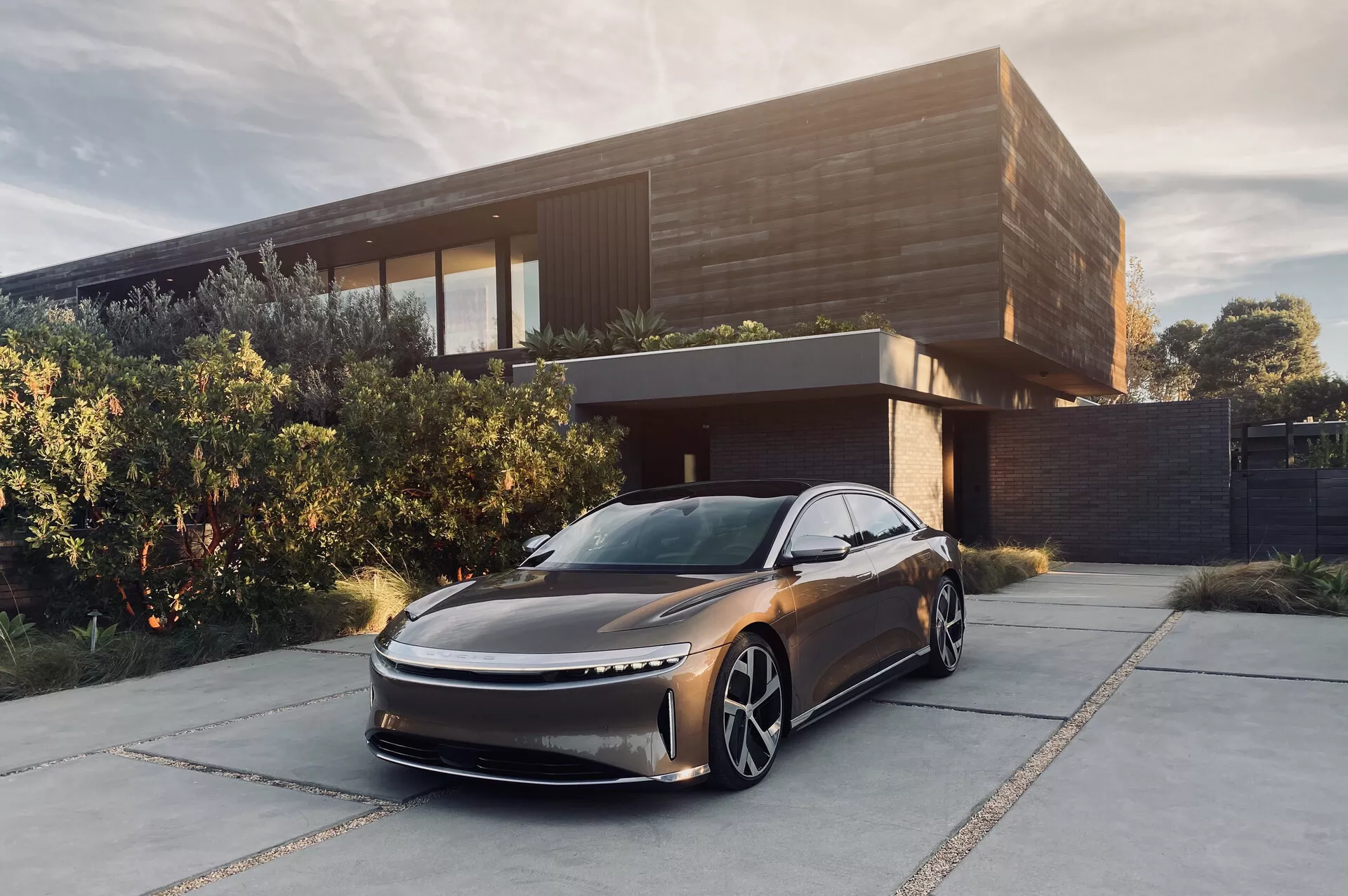 Obrazek przedstawiający model Lucid Air 2022, nowoczesny sedan, zaparkowany przed współczesnym domem. Zdjęcie z niskiego, przedniego trójćwiartkowego kąta, ukazujące jego smukły, aerodynamiczny design z wydłużoną maską i subtelną kratką. Widoczne są smukłe światła LED, duże charakterystyczne koła i brak widocznych bocznych lusterek sugerujących wykorzystanie kamer jako systemu wizyjnego. Metaliczny ciemny kolor i wyrafinowane otoczenie podkreślają jego luksusowy wygląd, sugerując, że jest to pojazd elektryczny skierowany do zamożnego segmentu rynku.