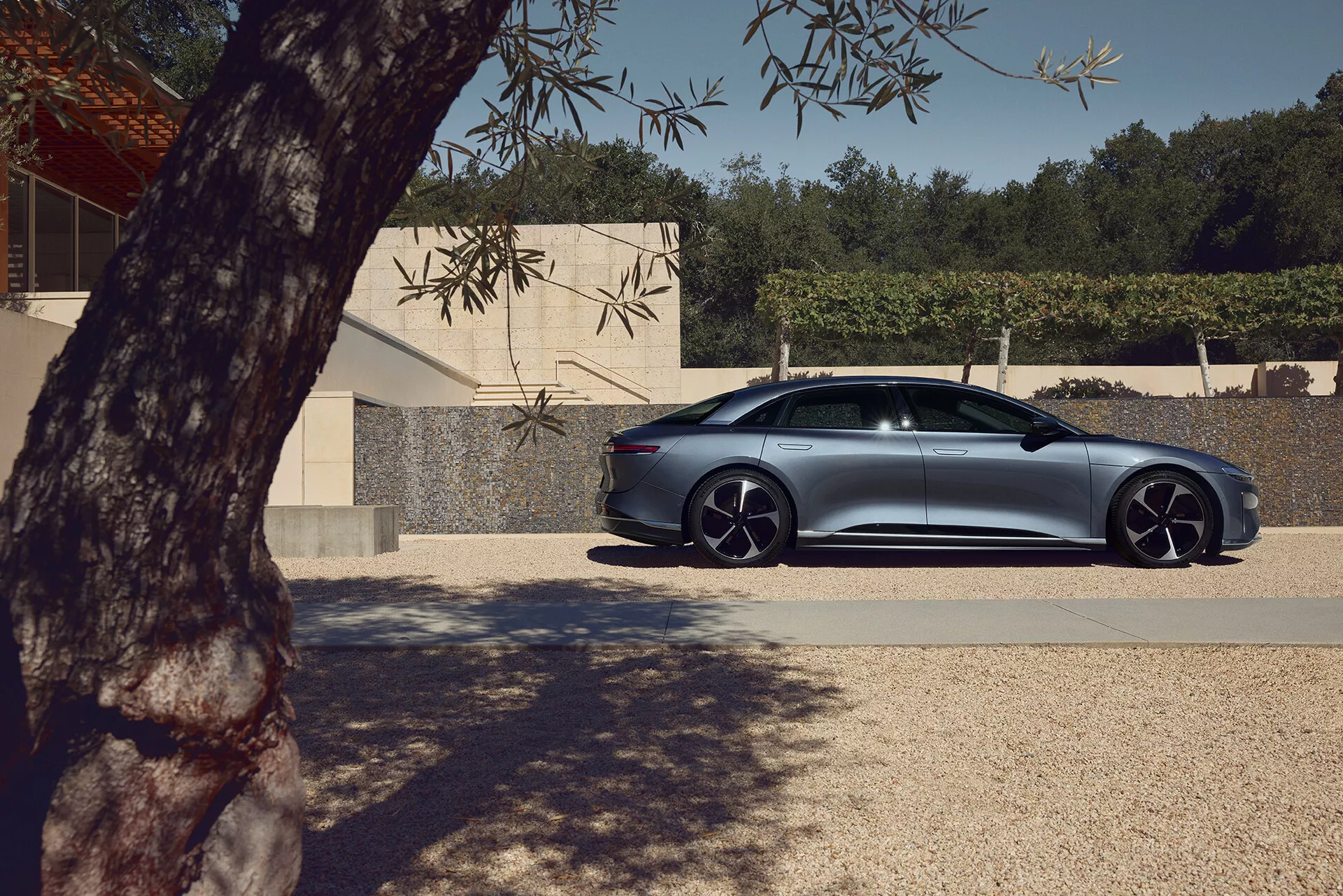 Widok z profilu na czterodrzwiowego sedana Lucid Air 2022 pierwszej generacji zaparkowanego na zewnątrz. Widoczny jest bok pasażera luksusowego, wysokowydajnego sedana, pokazujący gładkie linie nadwozia, design drzwi i okien oraz charakterystyczne aluminiowe koła z niskoprofilowymi oponami. Częściowo zacienione przez liście drzewa, z naturalnym światłem podkreślającym metaliczne wykończenie. Nowoczesny budynek w tle podkreśla elegancki, aerodynamiczny design samochodu z zaawansowanymi funkcjami, takimi jak uchwyty drzwi na równi z nadwoziem oraz aerodynamiczne lusterka boczne, mogące wskazywać na zintegrowane kierunkowskazy lub wykrywanie martwego punktu.