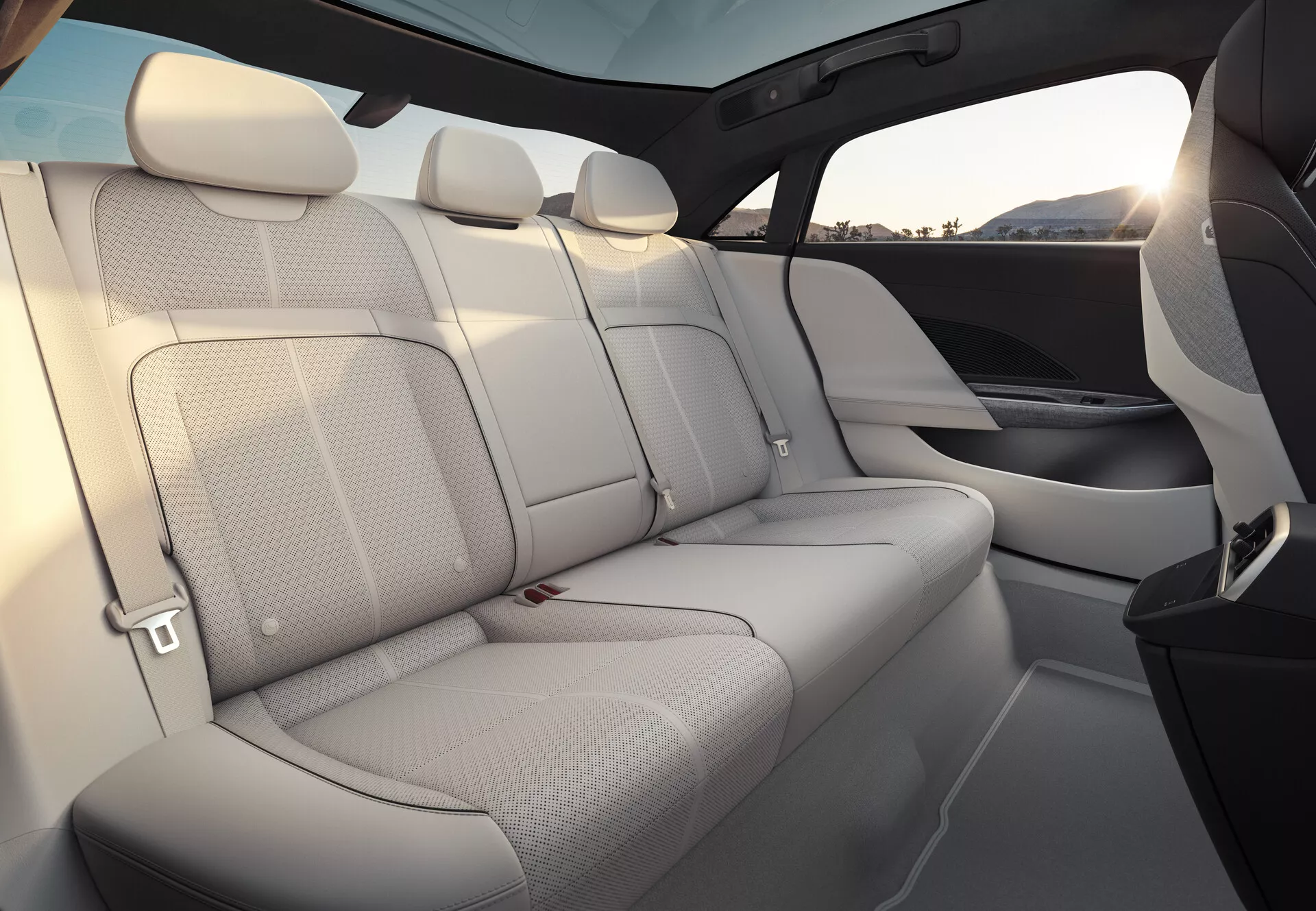 Widok wnętrza Lucid Air 2022, skoncentrowany na tylnych siedzeniach dla pasażerów. Siedzenia są wyściełane jasną skórą, oferując przewiewne i przestronne odczucie. Tylna ławka mieści trzech pasażerów z dopasowanymi zagłówkami i widocznymi pasami bezpieczeństwa. Panele drzwi harmonizują z materiałem i designem siedzeń, posiadające wbudowane kratki głośników. Wysoka linia okien i rozległy przyciemniony panoramiczny dach podnoszą poczucie prywatności i ochrony. Wykończenie wnętrza pokazuje mieszankę jasnej tapicerki i ciemniejszych elementów, prawdopodobnie wykonanych z materiałów wysokiej jakości.