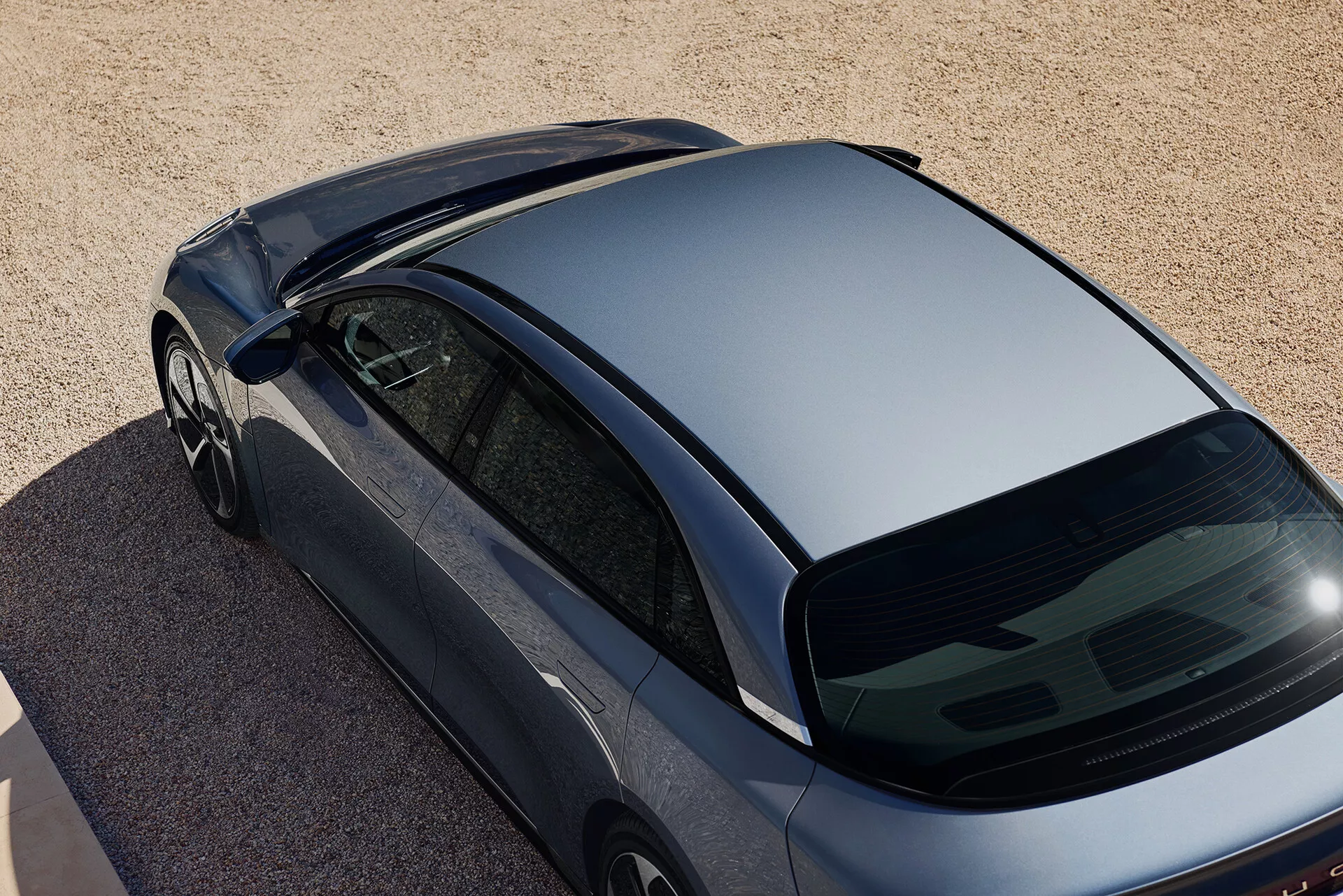 Ujęcie z góry, pod kątem, samochodu Lucid Air 2022 pierwszej generacji w ciemnym metalicznym kolorze, zaparkowanego na powierzchni z drobnych kamieni lub gruboziarnistego piasku. Zdjęcie uchwyca widok z góry, koncentrując się na opływowym dachu, stromo pochylonej tylnej szybie i części bagażnika. Cechy obejmują bezramkowe okna, opływowe lusterka boczne oraz brak akcesoriów dachowych i tylnej wycieraczki, co sugeruje aerodynamiczny i nowoczesny design.