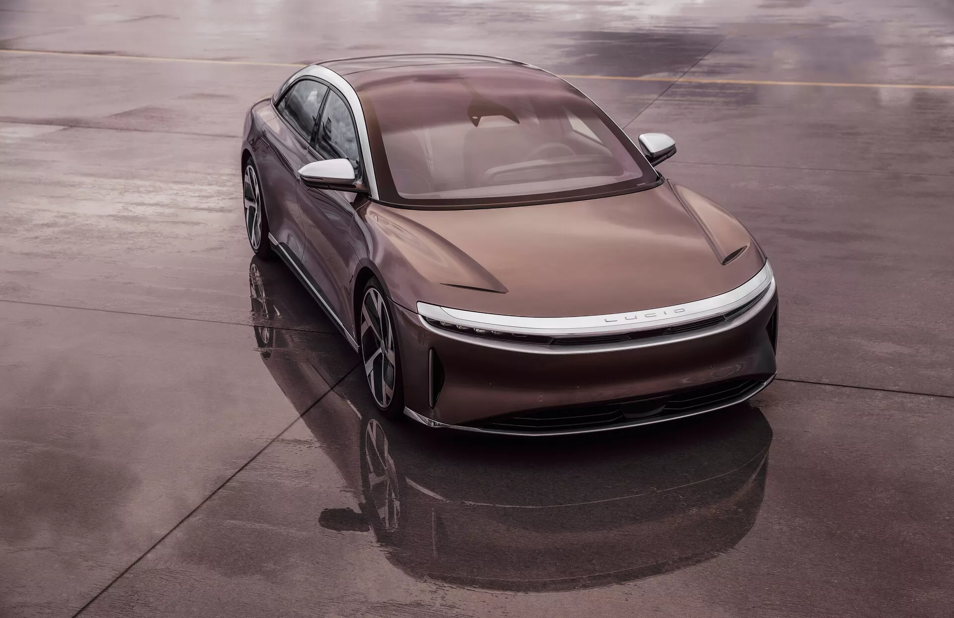 Zdjęcie pod wysokim kątem przedstawiające nowoczesny sedan Lucid Air 2022, 1. generacji, ustawiony pod skosem na odblaskowej powierzchni, takiej jak mokry asfalt, zapewniające kompleksowy widok na przednią lewą ćwiartkę pojazdu. Smukła, aerodynamiczna konstrukcja obejmuje gładkie krzywizny, cienkie reflektory LED i konturowaną maskę z wyraźnymi liniami. Dach fastback i wpuszczone szkło poprawiają wydajność aerodynamiczną. Elektryczny charakter samochodu sugeruje brak tradycyjnej kraty, zintegrowany system poboru powietrza i lekkie aerodynamiczne koła.