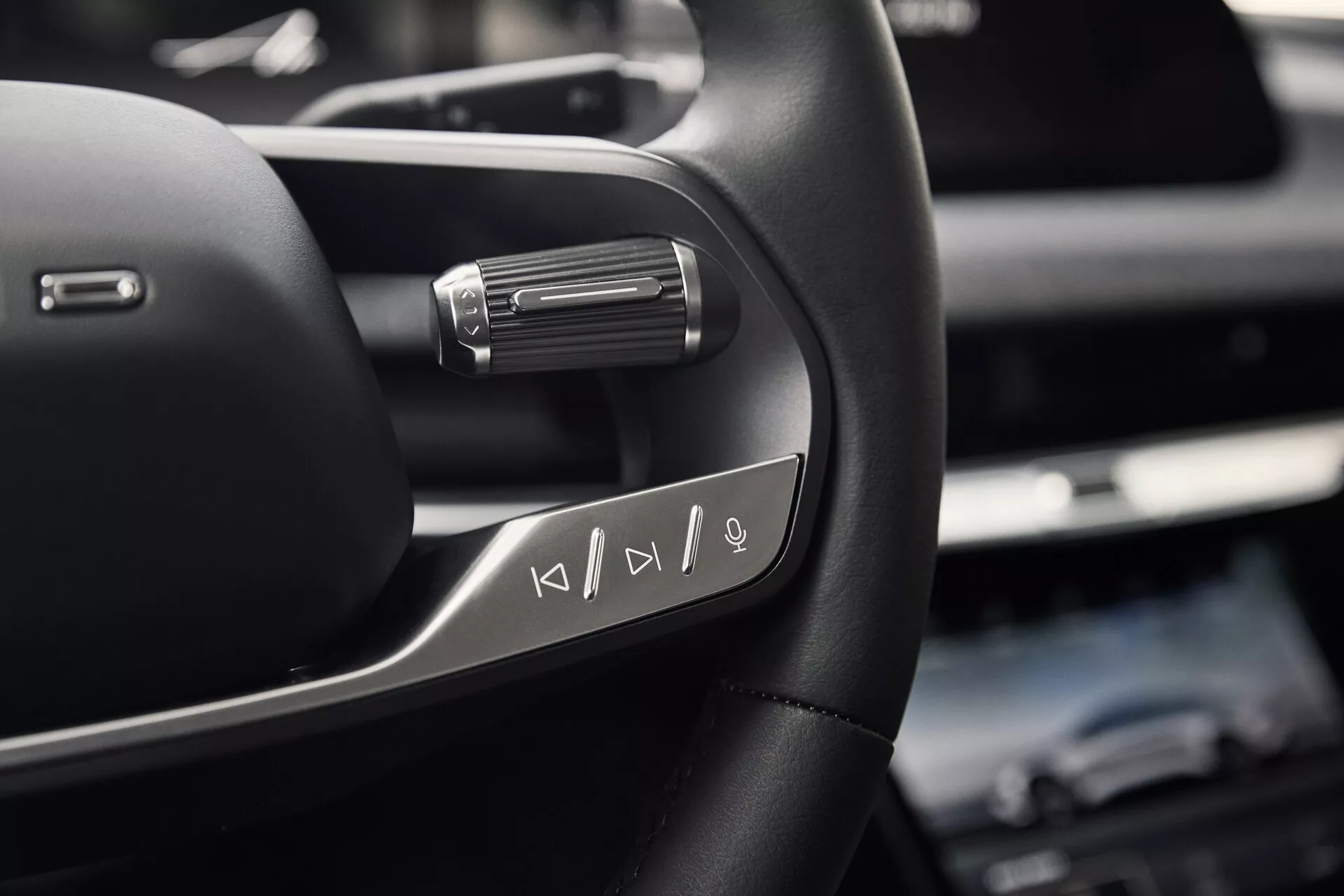 Zbliżenie lewej strony kierownicy Lucid Air 2022 z wielofunkcyjnymi przyciskami sterowania mediami. Zdjęcie wykonane z poniżej kierownicy na wysokości klatki piersiowej siedzącego kierowcy podkreśla wysokiej jakości skórzany materiał z metalowymi akcentami i łopatką za kierownicą, wskazując na zaawansowane funkcje jazdy.