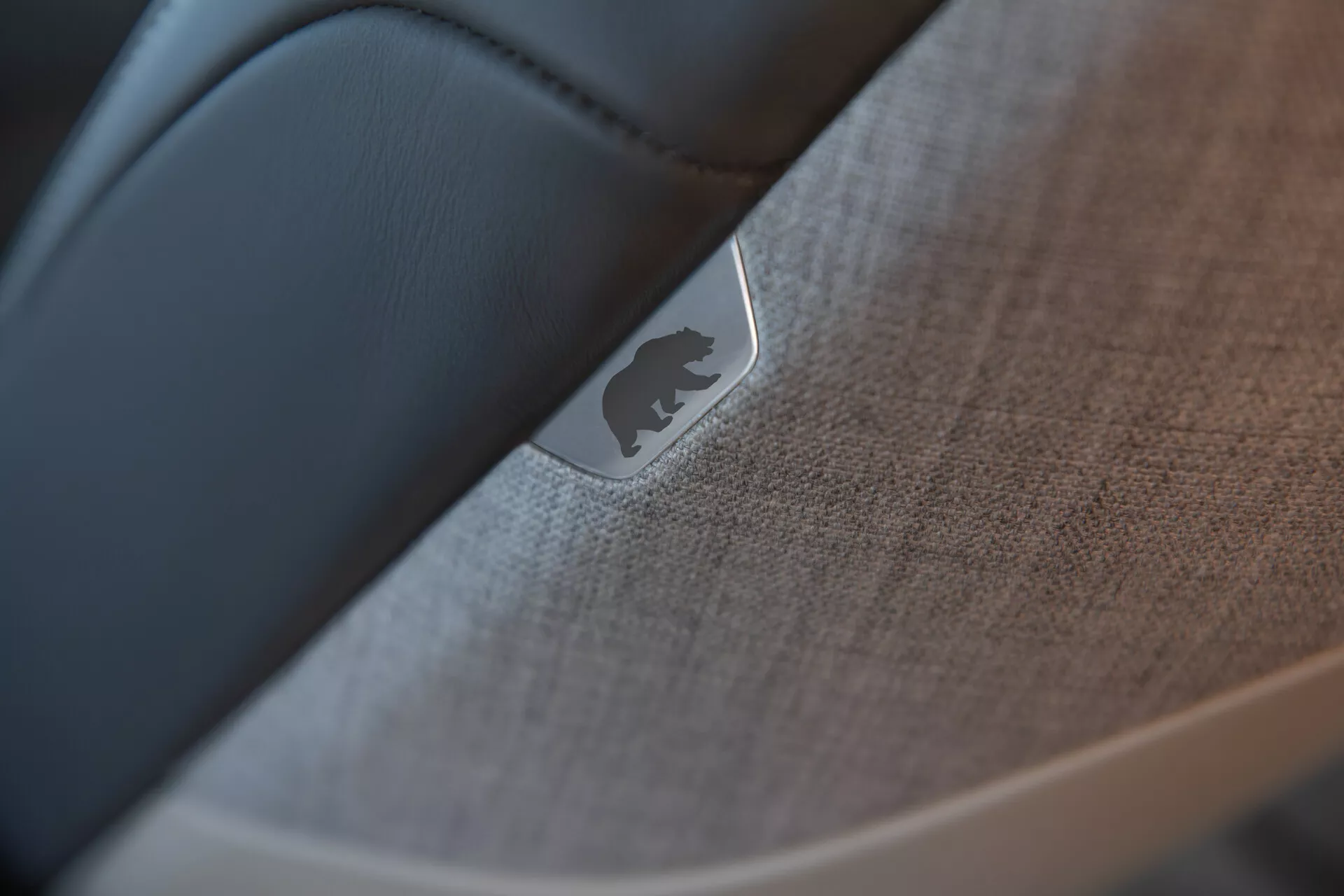 Zbliżenie, selektywne skupienie na wnętrzu samochodu Lucid Air 2022 (1. generacja) podkreślające szczegóły drzwi lub deski rozdzielczej. Refleksyjny metalowy emblemat lub odznaka jest wyraźnie widoczna, umieszczona na teksturowanej tkaninie lub miękkim plastiku z detalami szwów. Gładki, ciemniejszy materiał, prawdopodobnie czarna skóra, kontrastuje z teksturowanymi elementami, pokazując kunszt i wysoką jakość wnętrza samochodu.