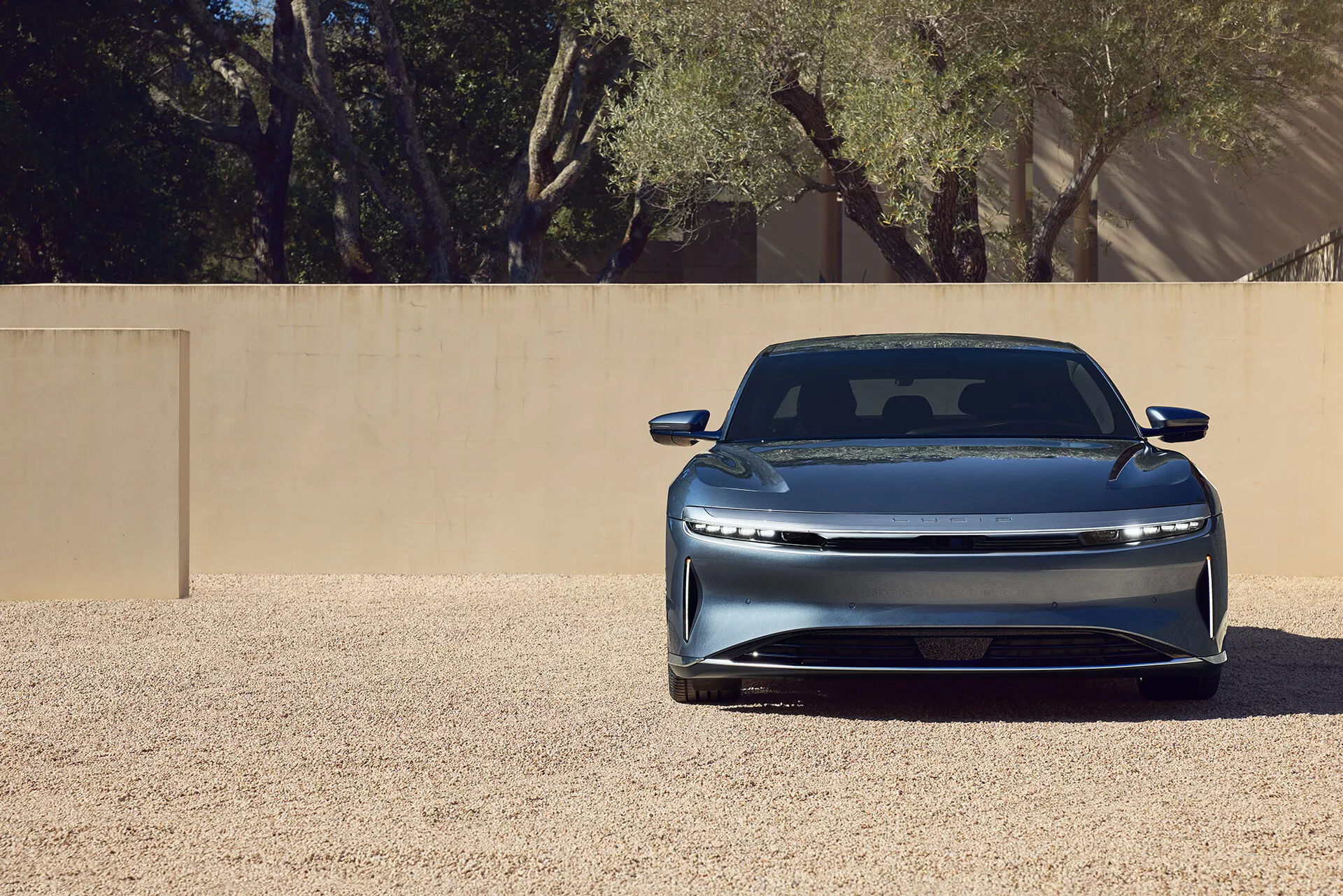 Zbliżenie przedniego ujęcia samochodu elektrycznego Lucid Air 2022 pierwszej generacji, podkreślające elegancki zamknięty grill, poziomo zorientowane reflektory LED, zintegrowany przedni zderzak, rzeźbioną maskę z linią konturu oraz metaliczne wykończenie lakieru. Samochód jest centralnie umieszczony na żwirowej powierzchni pod jasnym oświetleniem, co sugeruje zaawansowaną aerodynamikę i nowoczesny luksusowy design.