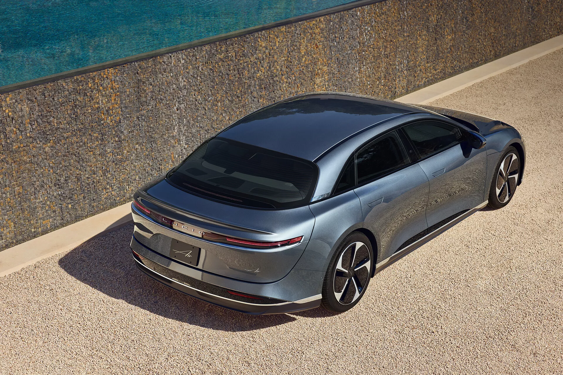 Zdjęcie z wysokiego kąta eleganckiego sedana Lucid Air 2022 widzianego z tylnej części pojazdu. Samochód ma sylwetkę coupé z opadającą linią dachu, wąskimi poziomymi światłami tylnymi i metalicznym wykończeniem lakieru. Otoczenie obejmuje morze i luksusową rezydencję, co dodaje poczucia wysokiej jakości. Minimalistyczny design zewnętrzny, duże koła wieloramienne i przyciemniane szyby sugerują nacisk na aerodynamikę i nowoczesną technologię, co może wskazywać na elektryczny układ napędowy.