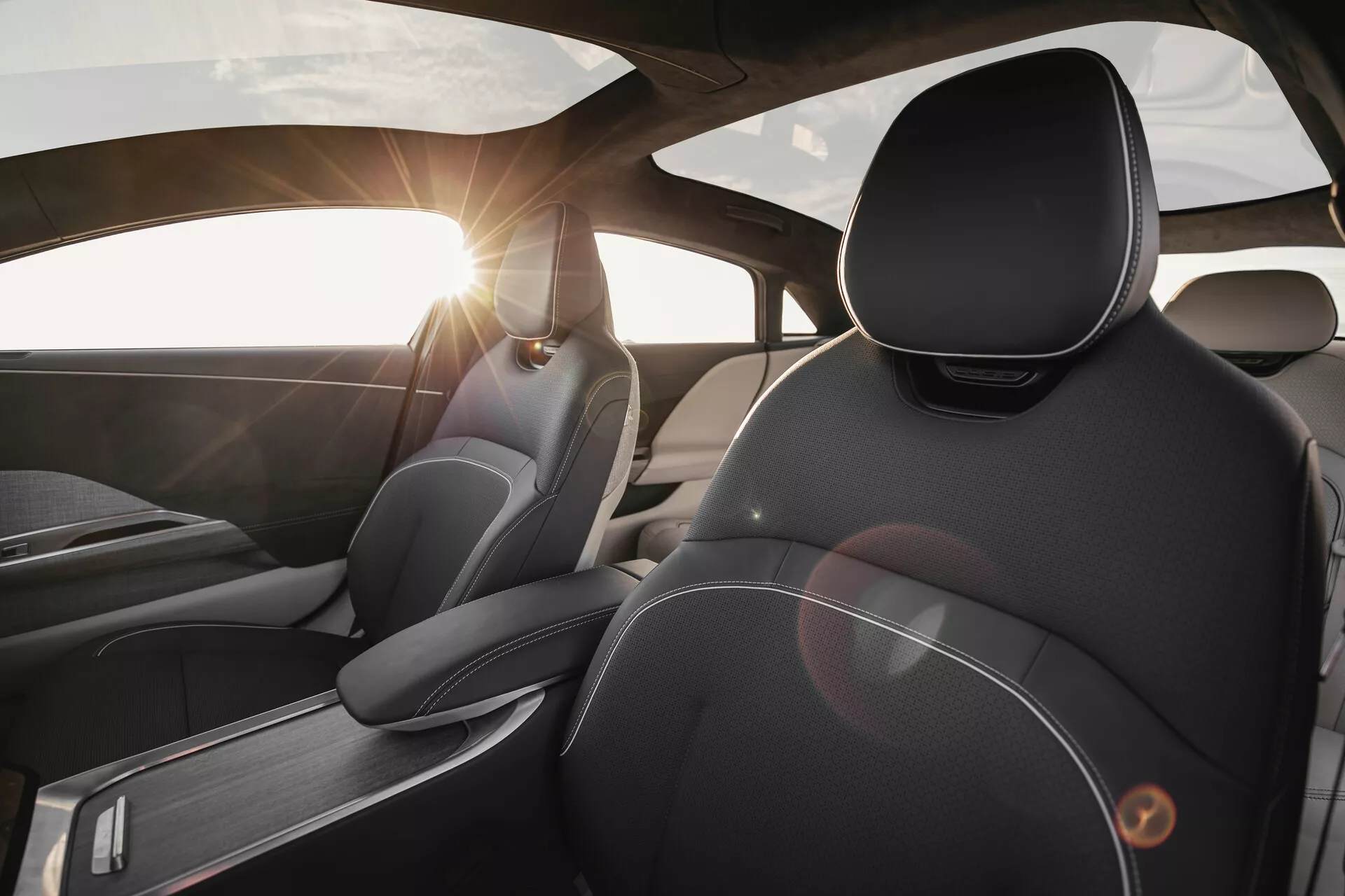 Widok wnętrza Lucid Air 2022 z perspektywy tylnego siedzenia, koncentrujący się na fotelu kierowcy. Skupiono się na wysokiej jakości materiałach i designie, z detalicznymi przeszyciami skórzanej tapicerki, ergonomicznie zaprojektowanych zagłówkach, akcentach z szczotkowanego metalu oraz efekcie flary słonecznej przez przednią szybę.