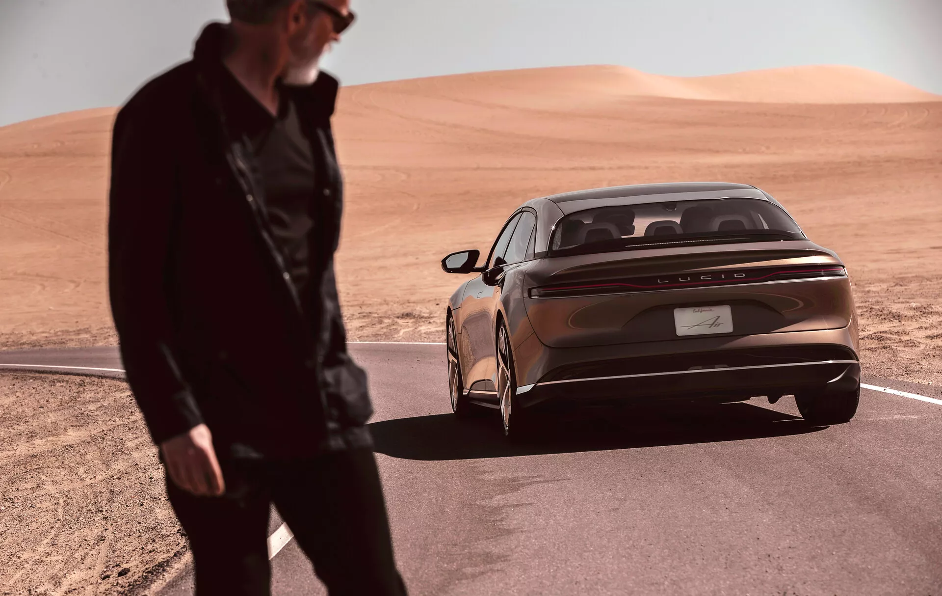 Elegancki i nowoczesny sedan Lucid Air 2022 (1. generacja) zaparkowany w krajobrazie pustynnym, uchwycony z niskiego kąta z naciskiem na tylną część wozu. Metaliczny, brązowy kolor nadwozia i wyraźne poziome tylne lampy podkreślają współczesny design samochodu. Odbijający światło pasek chromowany biegnie w poprzek szerokości bagażnika poniżej świateł. Znak producenta jest umieszczony centralnie na bagażniku. Zintegrowany tylny zderzak wyposażony jest w czujniki parkowania, co dodaje luksusowego wyglądu. Niewyraźna postać stoi na pierwszym planie, sugerując ruch lub aktywność w pobliżu pojazdu.