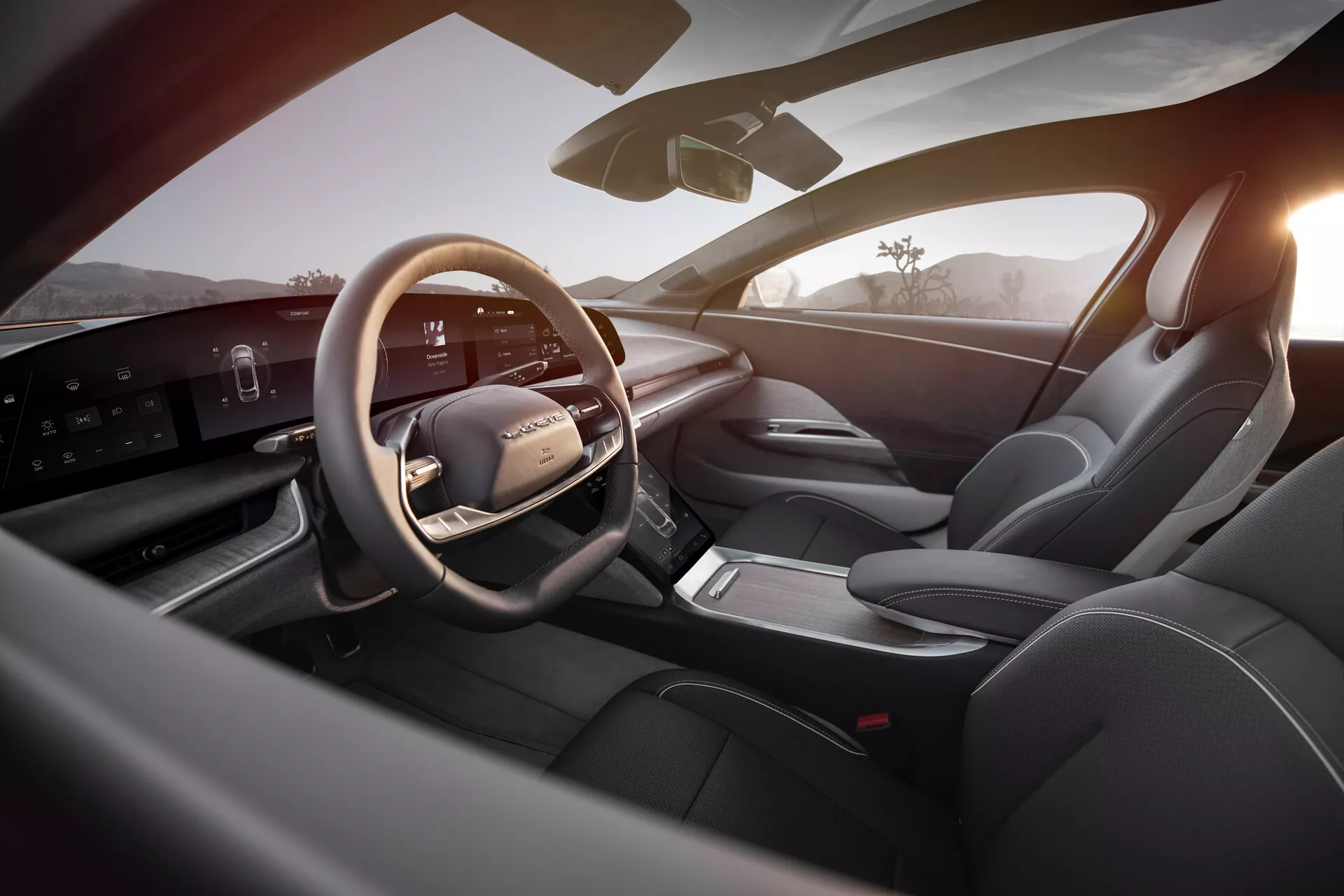 Widok wnętrza Lucid Air 2022 z perspektywy tylnego prawego siedzenia, ukazujący luksusowy i nowoczesny obszar kierowcy. Główne cechy to skórzana, wielofunkcyjna kierownica z zamontowanymi sterownikami, elegancki, poziomy cyfrowy wyświetlacz deski rozdzielczej, uproszczona konsola środkowa z elektronicznym selektorem biegów, sportowe fotele pokryte skórą, zaawansowane sterowniki panelu drzwi i dobra widoczność ogólna z bocznym lustrem i przednią szybą. Dobrze oświetlone wnętrze podkreśla nowoczesną technologię i luksusowy design, uwydatniony ciepłym, zewnętrznym światłem sugerującym wschód lub zachód słońca.