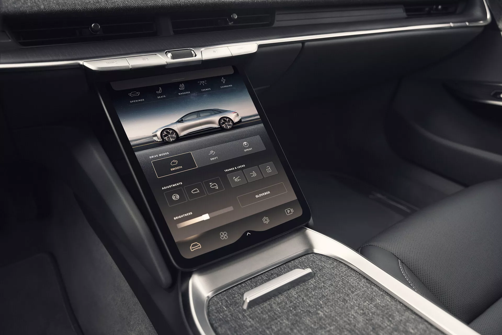 Widok wnętrza Lucid Air 2022 (1. generacja) przedstawiający konsolę środkową z pionowo zamontowanym zintegrowanym ekranem dotykowym, zawierającym ikony nawigacji, telefonu, mediów, klimatyzacji i ustawień pojazdu. Zdjęcie zrobione z lekko pochylonej w lewo perspektywy uwydatnia zaawansowane funkcje technologiczne z otworami wentylacyjnymi nad ekranem, lśniącymi okuciami z chromu lub metalu, elegancką deską rozdzielczą i mieszanką skórzanych oraz metalowych wykończeń. Obrazek szczegółowo ukazuje również małą półeczkę pod ekranem, metalowy wybierak biegów oraz skórzane podłokietniki i boki siedzeń, podkreślając luksusowe i wygodne wnętrze.