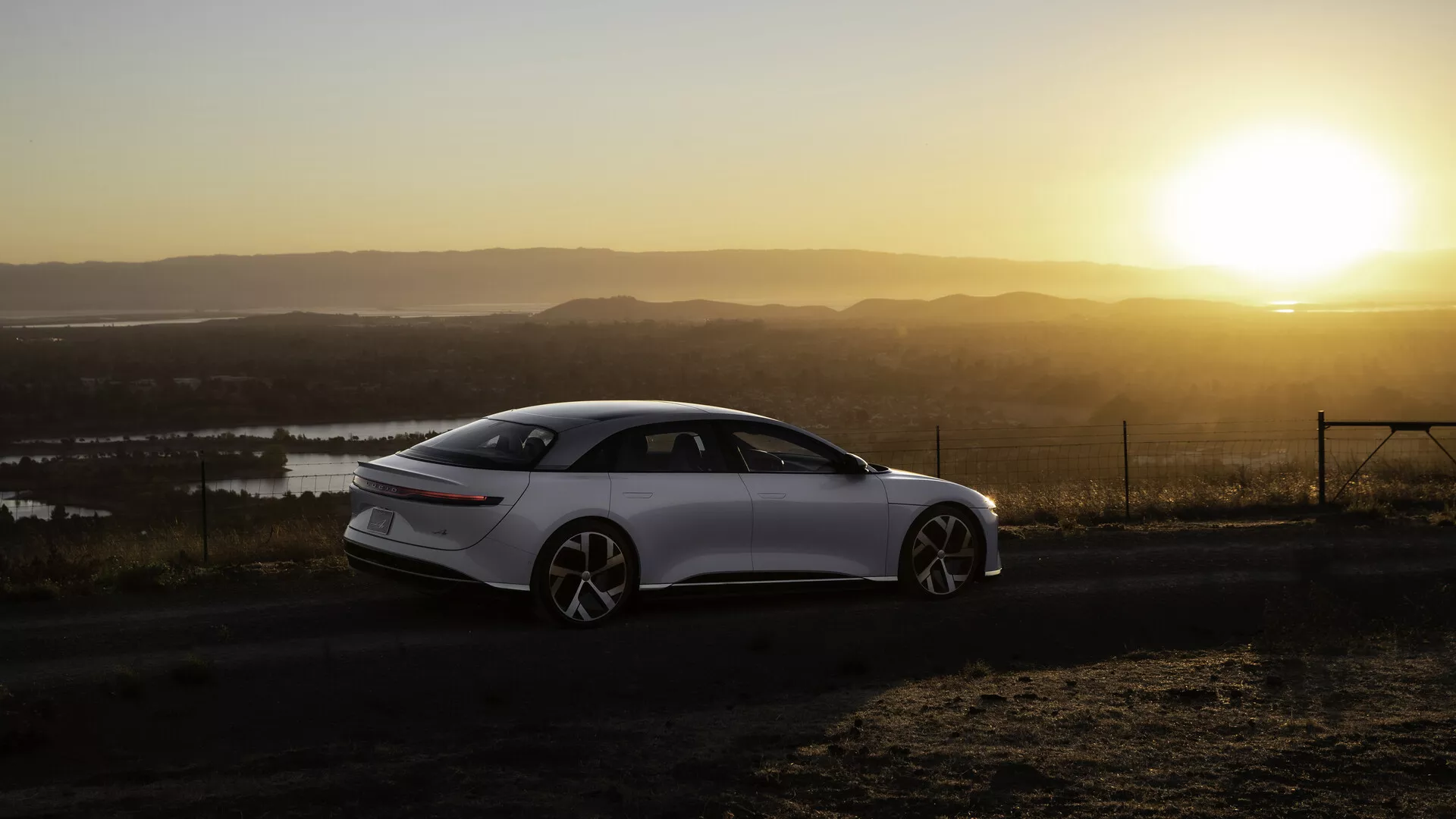 Lucid Air 2022 (1. Generacja) sfotografowany z bocznego tylnego kąta podczas wschodu lub zachodu słońca z ciepłym słońcem w tle, umieszczony po lewej stronie kadru. Elegancki, nowoczesny sedan o czterech drzwiach ma gładkie linie, duże wieloszprychowe koła i chowane klamki drzwi. Krajobraz obejmuje zbiorniki wodne i punkt widokowy na wysokości. Tylne światła są wąskie i poziomo wydłużone, a światło słoneczne tworzy efekt ciepłej poświaty i blasku na scenie.