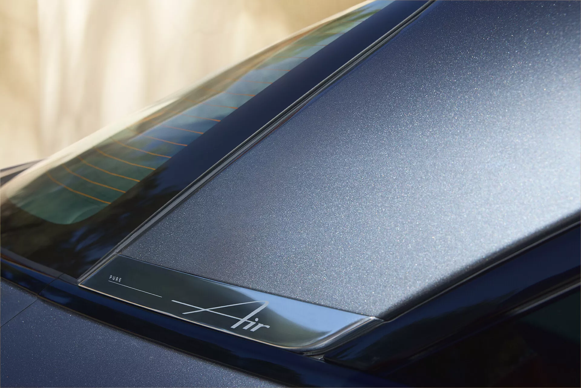 Zbliżenie tylnej części przy dachu samochodu Lucid Air 2022 (1. generacja), podkreślające ciemnoniebieskie błyszczące wykończenie i eleganckie linie aerodynamiczne. Zawiera podświetlany pasek z napisem 'Air', podkreślający luksusowy design i możliwą zaawansowaną technologię.
