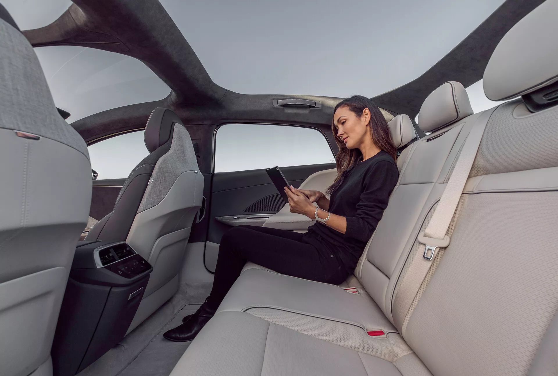 Wnętrze tylnej części kabiny Lucid Air 2022, podkreślające przestronność i luksus kabiny z jasnoszarymi, profilowanymi skórzanymi siedzeniami. Obraz uchwycony w szerokokątnym ujęciu z prawej strony, przedstawia panoramiczny dach rozciągający się nad tylną przestrzenią, ergonomicznie zaprojektowane siedzenia i złożony środkowy podłokietnik z uchwytami na kubki. Kobieta siedząca wygodnie korzysta z tabletu, wskazując na potencjalne funkcje łączności. Eleganckie i minimalistyczne wnętrze z czerwonymi przyciskami zwalniającymi pasy bezpieczeństwa i poziomymi liniami na desce rozdzielczej i panelach drzwiowych, wzmacniające poczucie szerokości i otwartości.