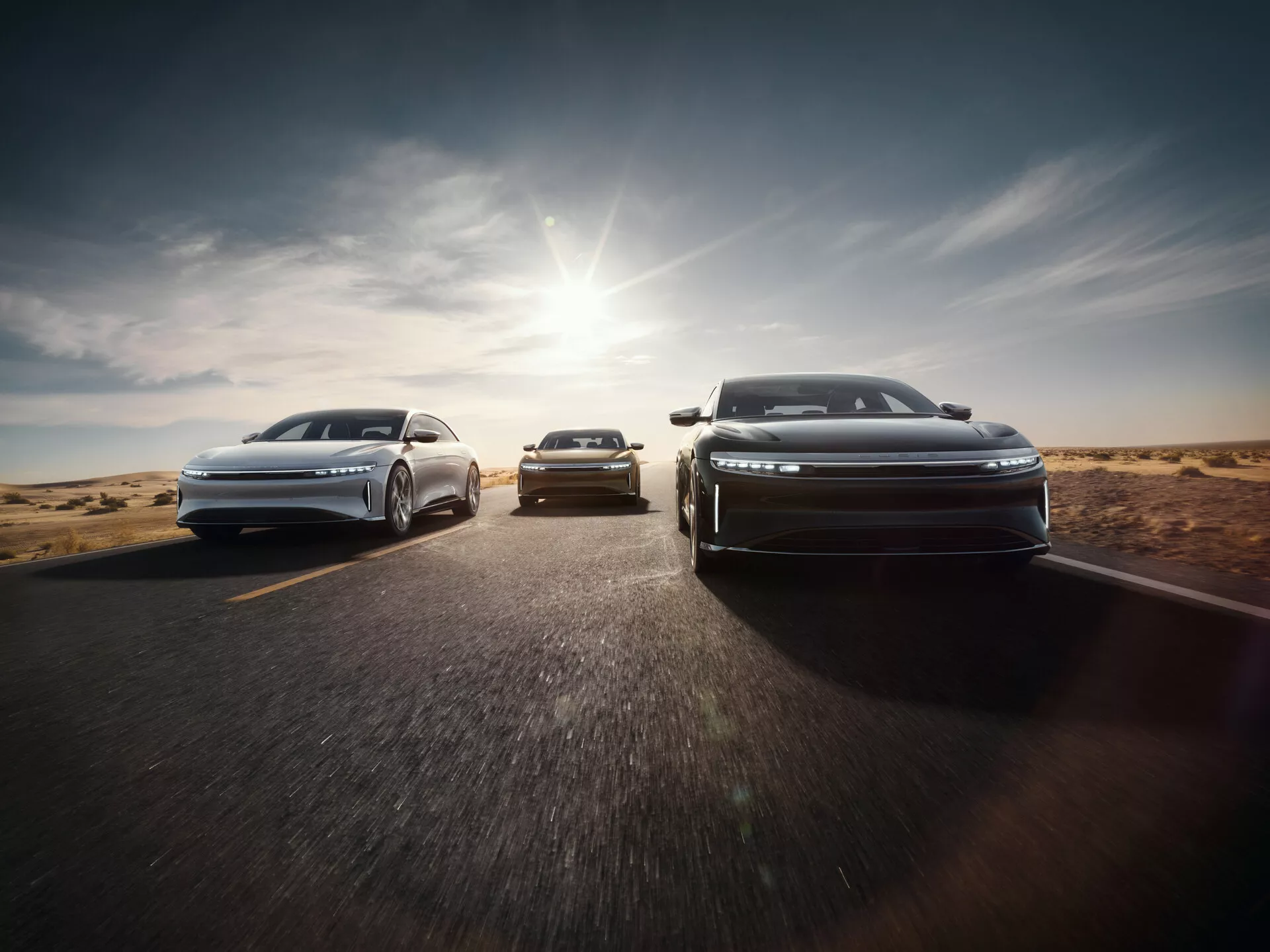 Widok z przodu Lucid Air 2022 z aerodynamicznym designem i reflektorami LED w formacji schodkowej na drodze asfaltowej z pustynnym tłem, światło słoneczne rzucające ciepły blask