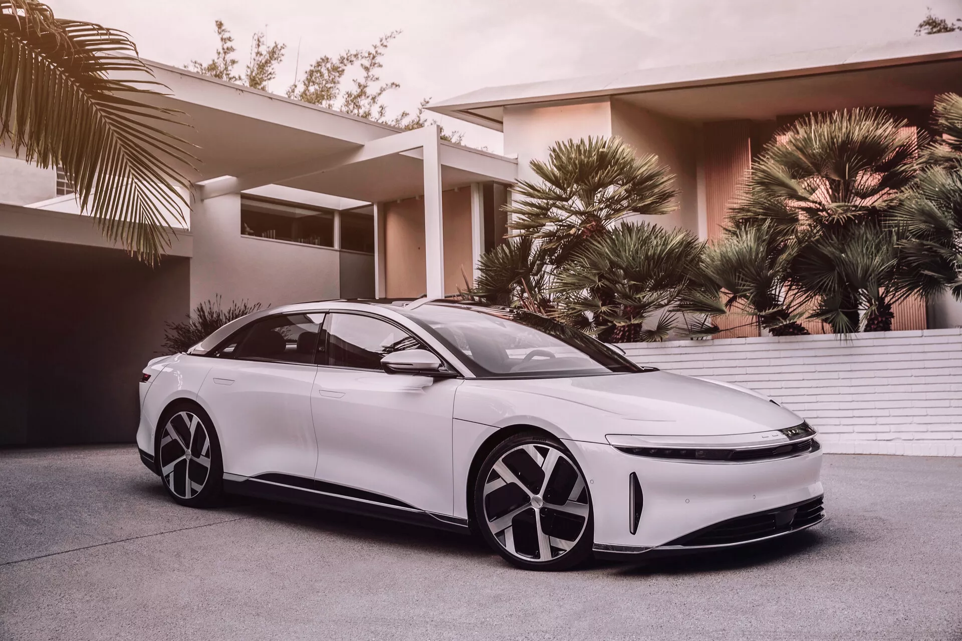 Obraz przedstawia Lucid Air 2022 pierwszej generacji, nowoczesny, elegancki biały sedan o niskiej, szerokiej sylwetce i linii dachu przypominającej coupe. Zdjęcie wykonane z niskiego kąta akcentuje aerodynamiczny design, duże, charakterystyczne felgi, reflektory LED i opływowe lusterka boczne, sugerujące zaawansowane technologie wspomagania kierowcy. Minimalistyczna przednia kratka wskazuje na technologie napędu elektrycznego lub hybrydowego. Futurystyczny design felg posiada aerodynamiczne osłony, a wygląd pojazdu poprawiają przyciemniane szyby, bezramowe drzwi i eleganckie, wpuszczane klamki. Częściowy widok panoramatycznego dachu sugeruje luksusowe, przestronne wnętrze. Tło pokazuje luksusową dzielnicę mieszkaniową z palmami, sugerując, że samochód jest przeznaczony dla zamożnych konsumentów.