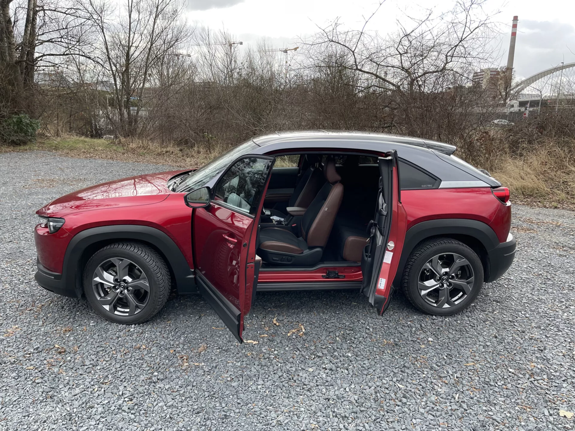 Czerwone SUV Mazda MX-30 z czterema drzwiami z 2020 roku w profilu bocznym na żwirze z otwartymi drzwiami, alufelgami o wielu szprychach, przyciemnianymi oknami i dachem w kontrastowym kolorze na tle przemysłowym i z zielenią.