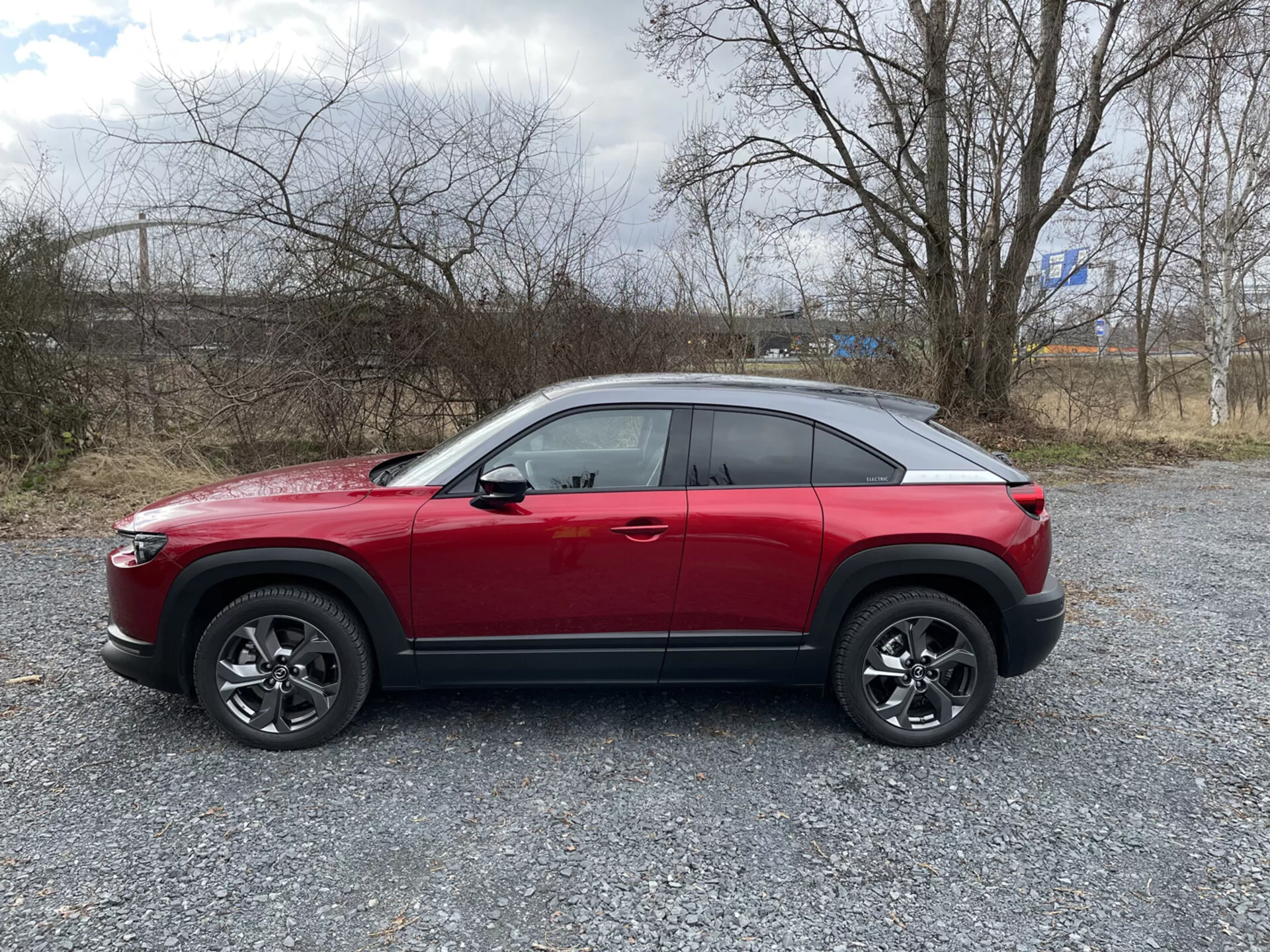 Profil boczny czerwonego crossovera SUV Mazda MX-30 2020 na żwirze ze sporą roślinnością i pochmurnym niebem, prezentujący wysoką linię pasa, dach opadający ku tyłowi na wzór coupé, dynamiczną linię charakterystyczną, wieloramienne felgi z czarnymi osłonami i możliwe zaawansowane funkcje technologiczne takie jak systemy ADAS, oświetlenie LED, system bezkluczykowego dostępu, system monitorowania ciśnienia w oponach TPMS i system infotainment.