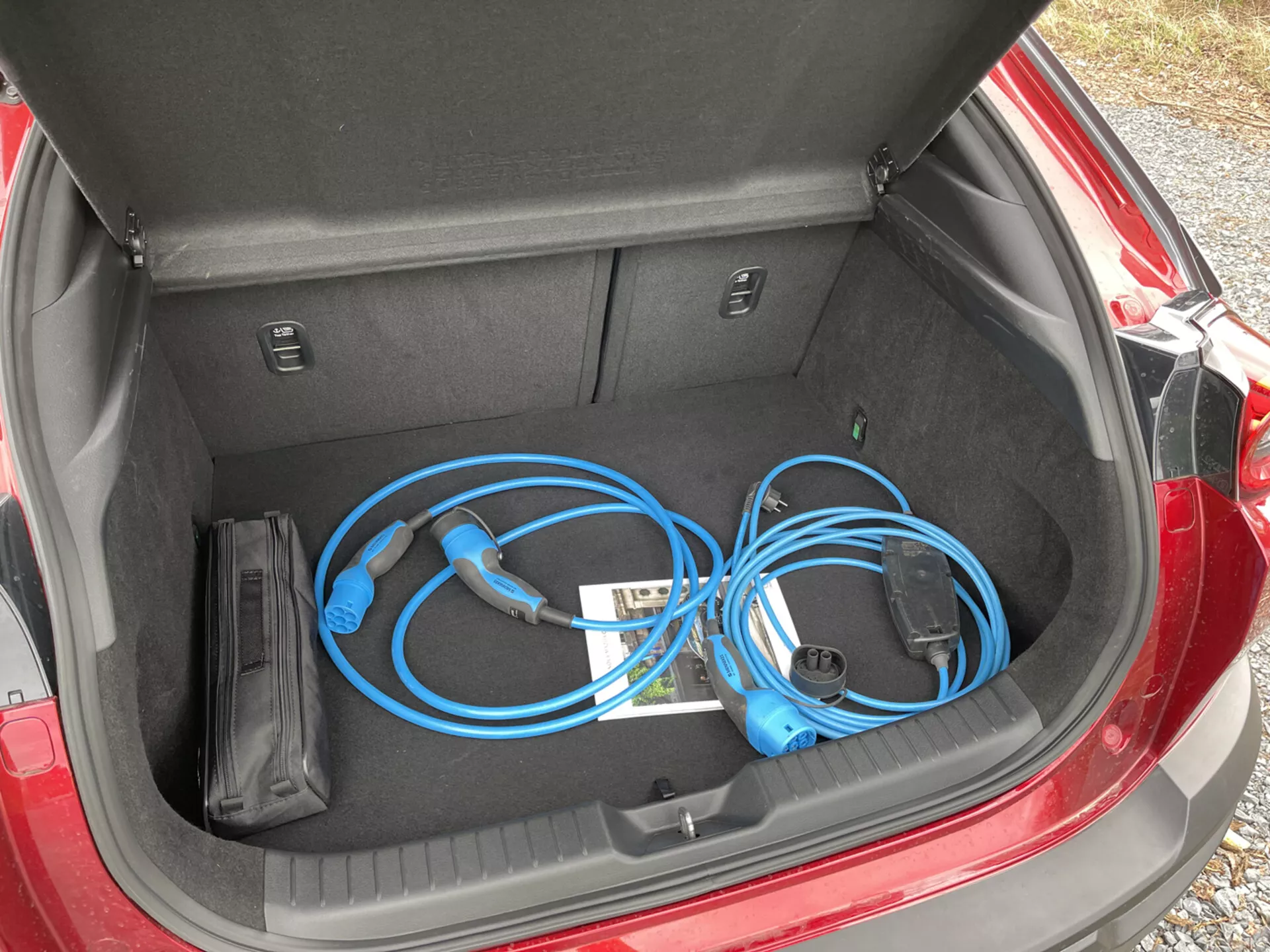 Otwarty bagażnik Mazdy MX-30 2020 z przenośnym kablem ładowania do pojazdów elektrycznych, z bliska, widok z góry od lewej strony, prezentujący czystą i płaską przestrzeń ładunkową wraz z wewnętrznymi przegrodami.