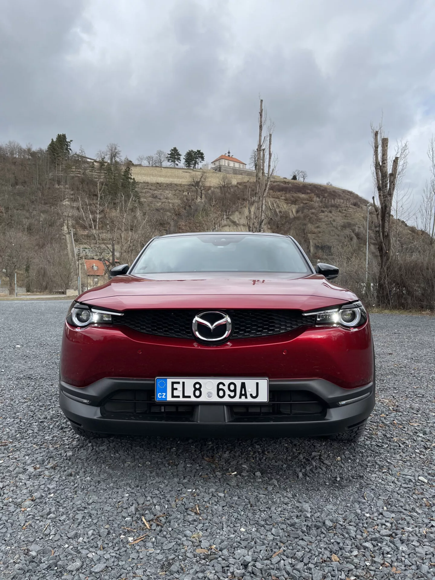 Przedni widok na czerwone Mazda MX-30 z 2020 roku, podkreślający kratkę, zderzak i reflektory LED samochodu na żwirowej powierzchni na tle pochmurnego nieba z zielenią i budynkiem