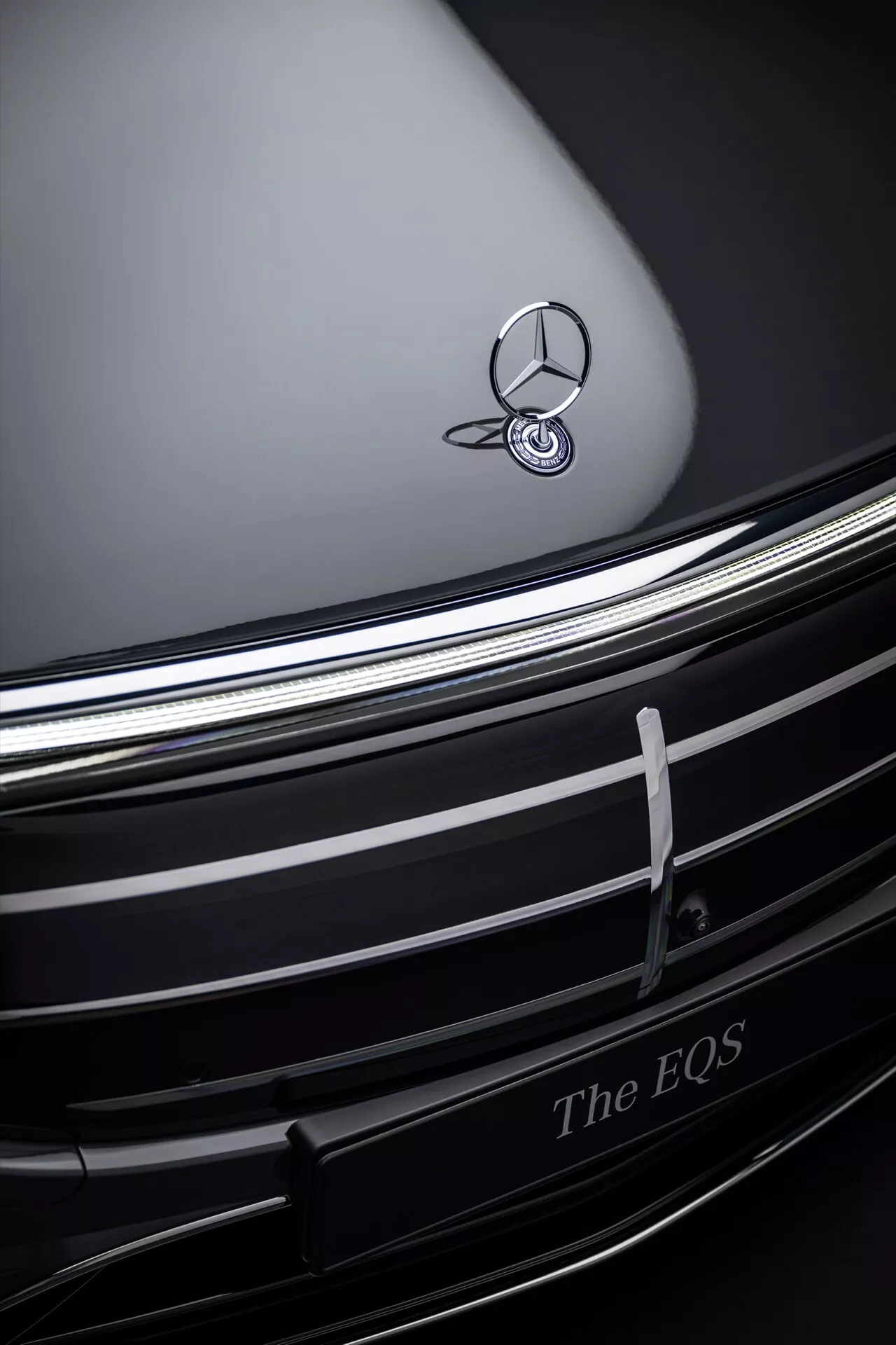 Zbliżenie na przedni grill i emblemat Mercedes-Benz EQS 2024, prezentujące chromowany emblemat na ciemnej konturowanej masce, z eleganckimi poziomymi chromowanymi żaluzjami i napisem 'The EQS', co wskazuje na design luksusowego pojazdu elektrycznego