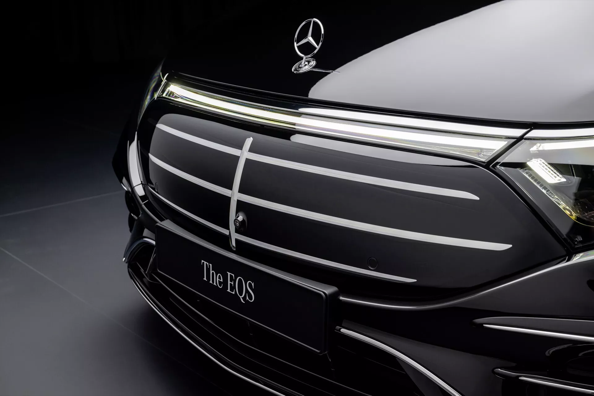 Zbliżenie na przednią część modelu Mercedes-Benz EQS 2024 po liftingu, prezentujące obszar grilla i emblemat marki na wyrafinowanym ciemnym tle, podkreślające elegancki design i elektryczne cechy luksusowe pojazdu