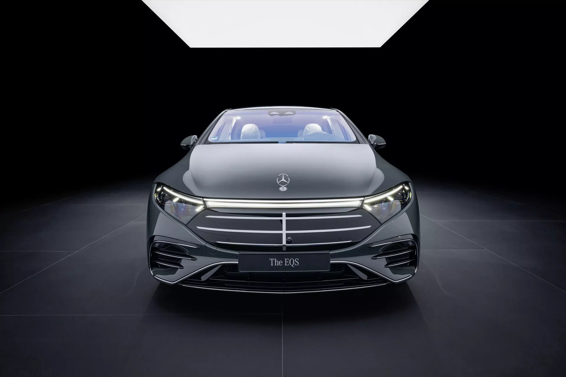 Przedni widok Mercedes-Benz EQS 2024 (297-Facelift) z wyrazistą atrapą z emblematem, smukłymi reflektorami LED i aerodynamicznym designem maski, wystawiony pod dramatycznym światłem reflektorów na targach motoryzacyjnych