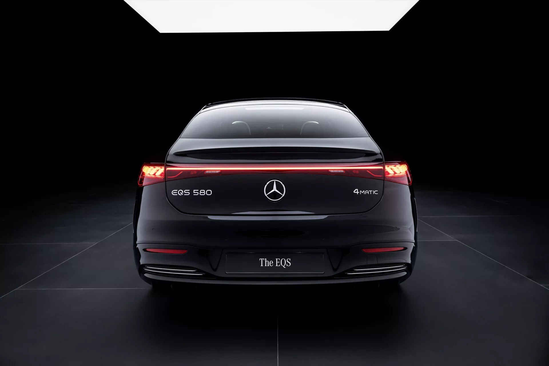 Tylny widok Mercedes-Benz EQS 580 4MATIC 2024 z eksponowaną szeroką postawą, podświetlonymi lampami tylnymi z poziomym pasem świetlnym, oznaczeniem modelu i zaawansowanymi technologiami bezpieczeństwa na ciemnym, gradientowym tle podkreślającym jego aerodynamikę i elektryczny design pojazdu.