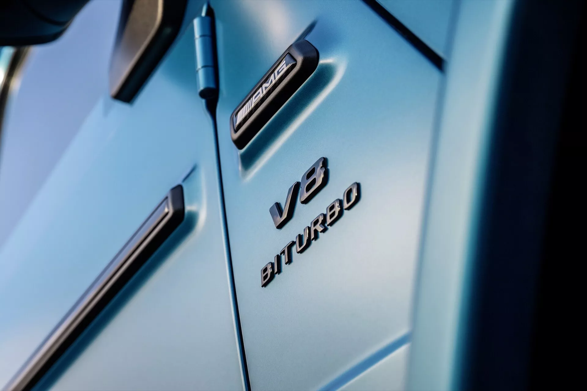 Zbliżenie na przedni błotnik Mercedes-Benz G-Klasse 2024 z emblematami V8 BITURBO i 4MATIC, ukazujące luksusowe linie projektowe i błyszczące, jasnoniebieskie wykończenie